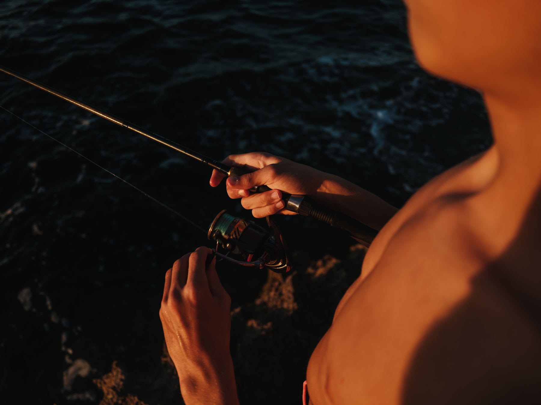 Over the shoulder view of a shirtless man holding a fishing rod and reel in the warm light of sunset over dark water.