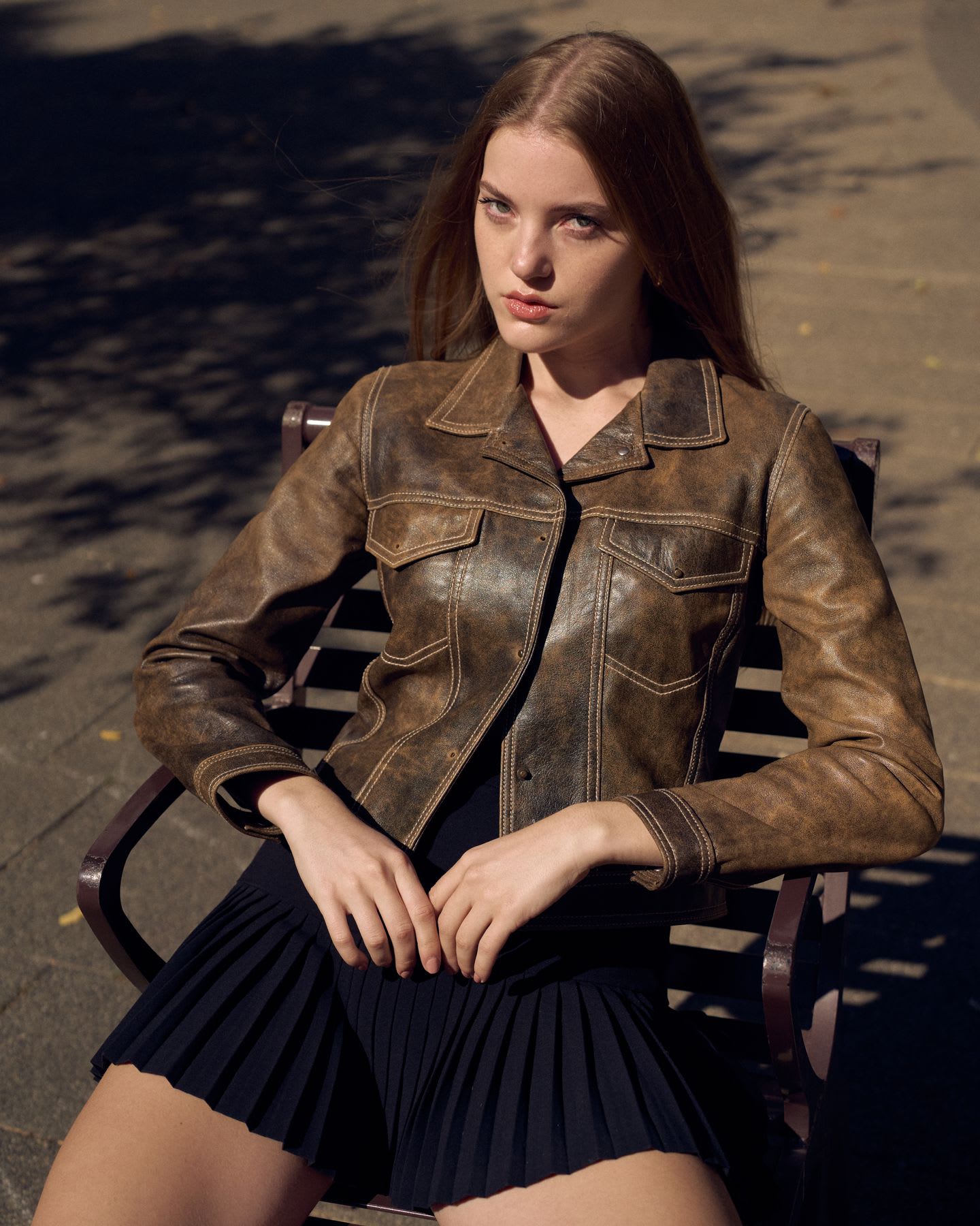 A young woman wearing a textured jacket and black pleated skirt sits on an outdoor bench in bright sunlight, looking directly at the camera.