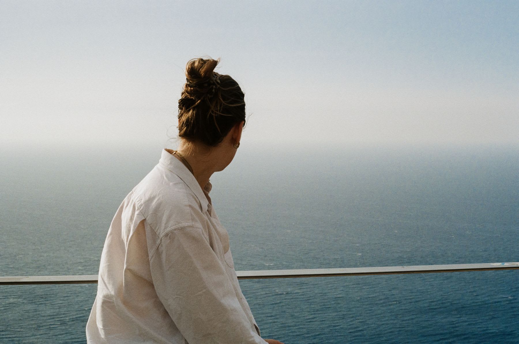 A woman with her hair in a bun sits and looks out at the expansive, calm ocean stretching to the horizon.