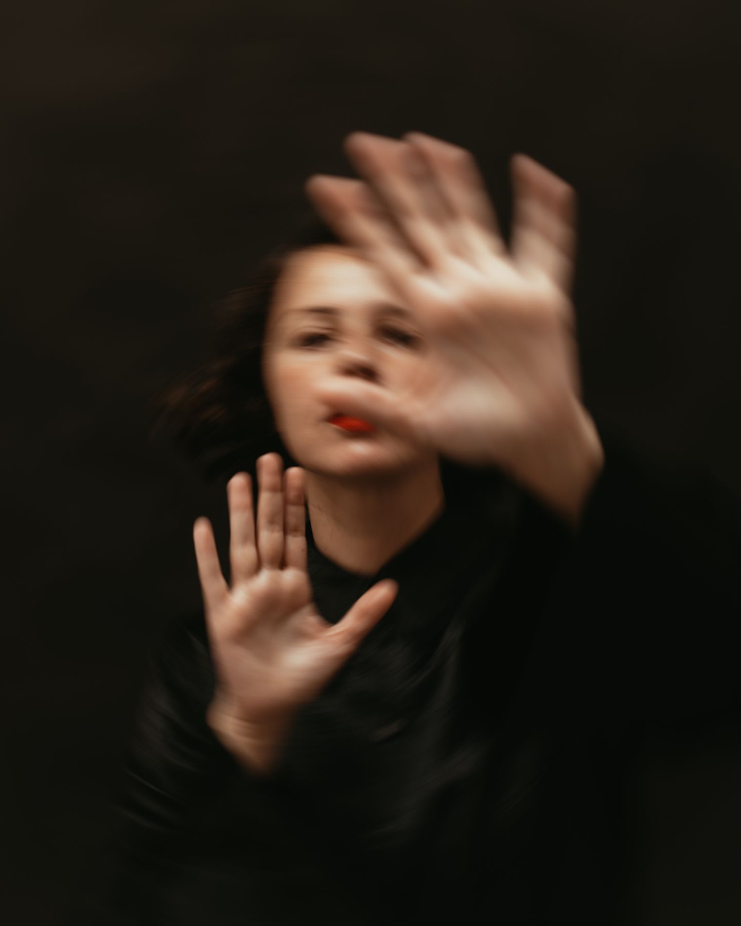 A blurred person with their hands raised, as if covering their face, against a dark background.