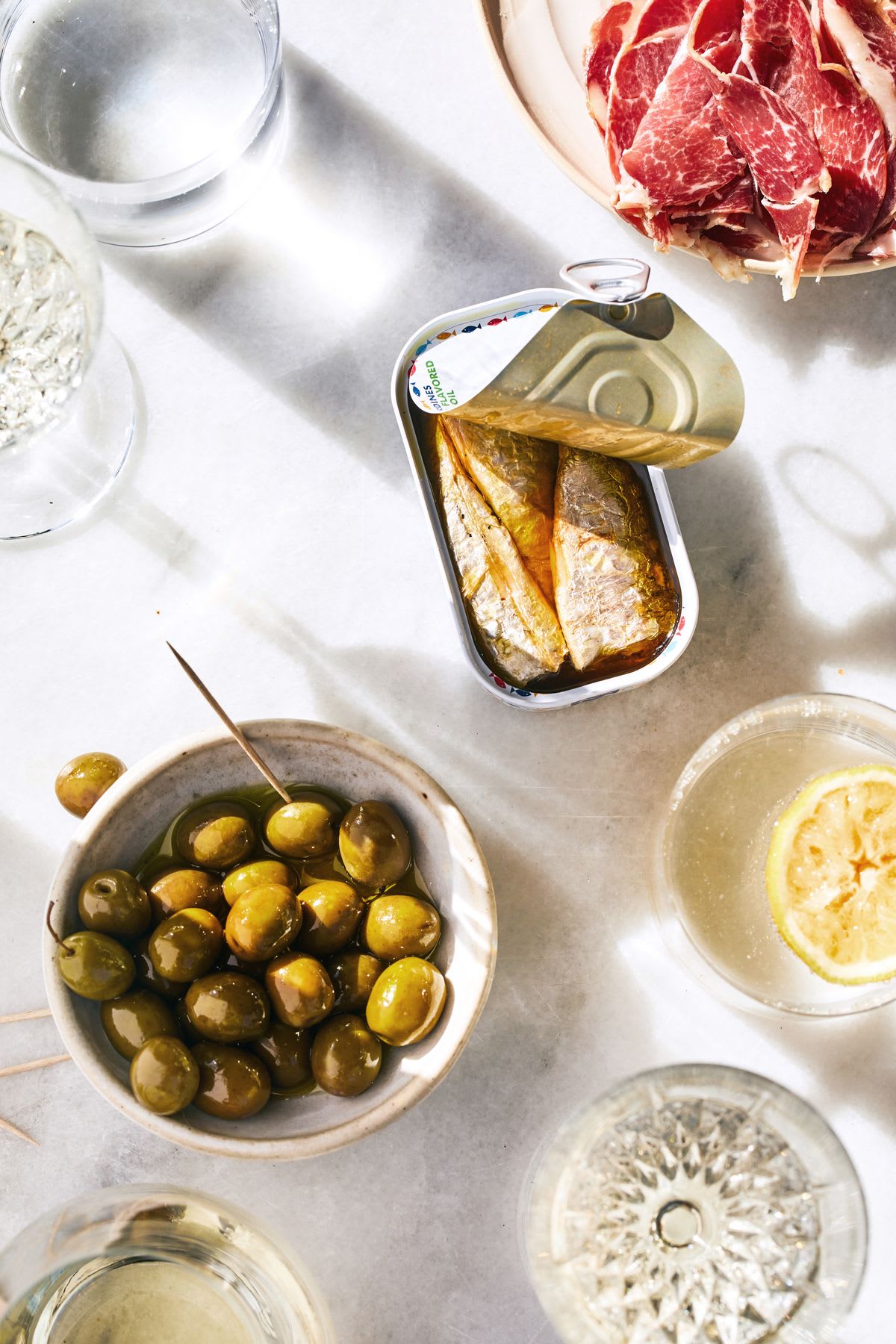 Table with white tablecloth and olive bowls, raw meat and a can of sardines