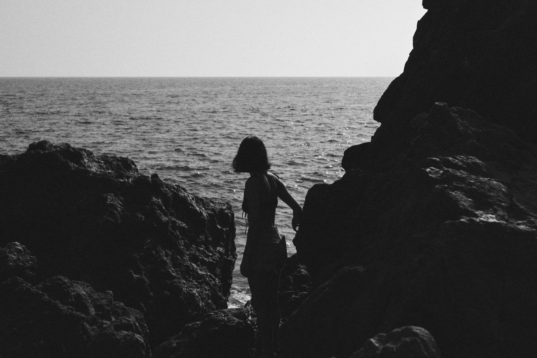 A girl stands between some jagged rocks overlooking the ocean.