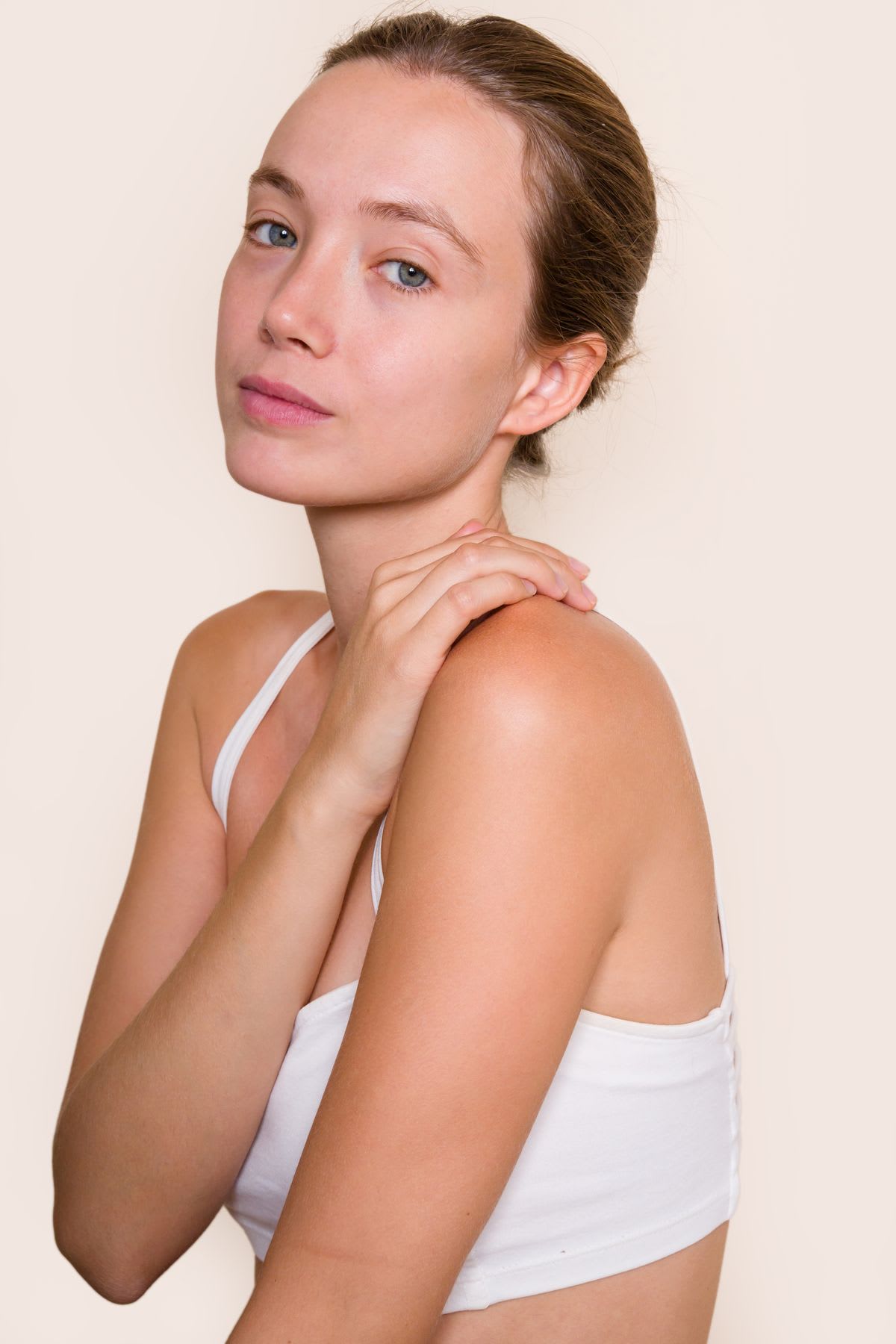 A woman wearing a white top, with her hair caught behind her head, holding her own shoulder.