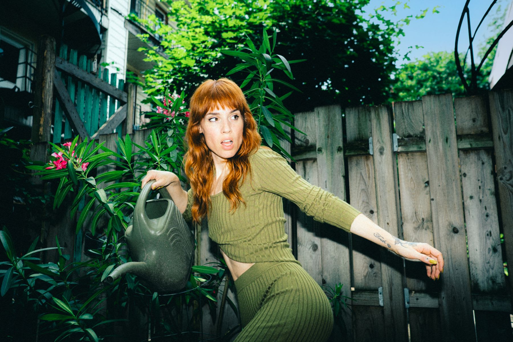 A stylish woman with red hair makes a surprised expression while holding a watering can in the middle of a lush, green backyard garden.