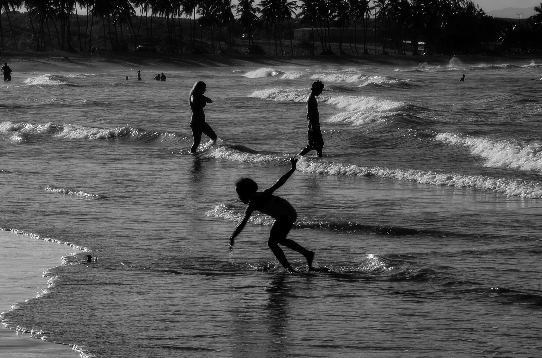 Children are playing in the shallow waves of the beach, silhouetted against the setting sun, with palm trees lining the distant shore.