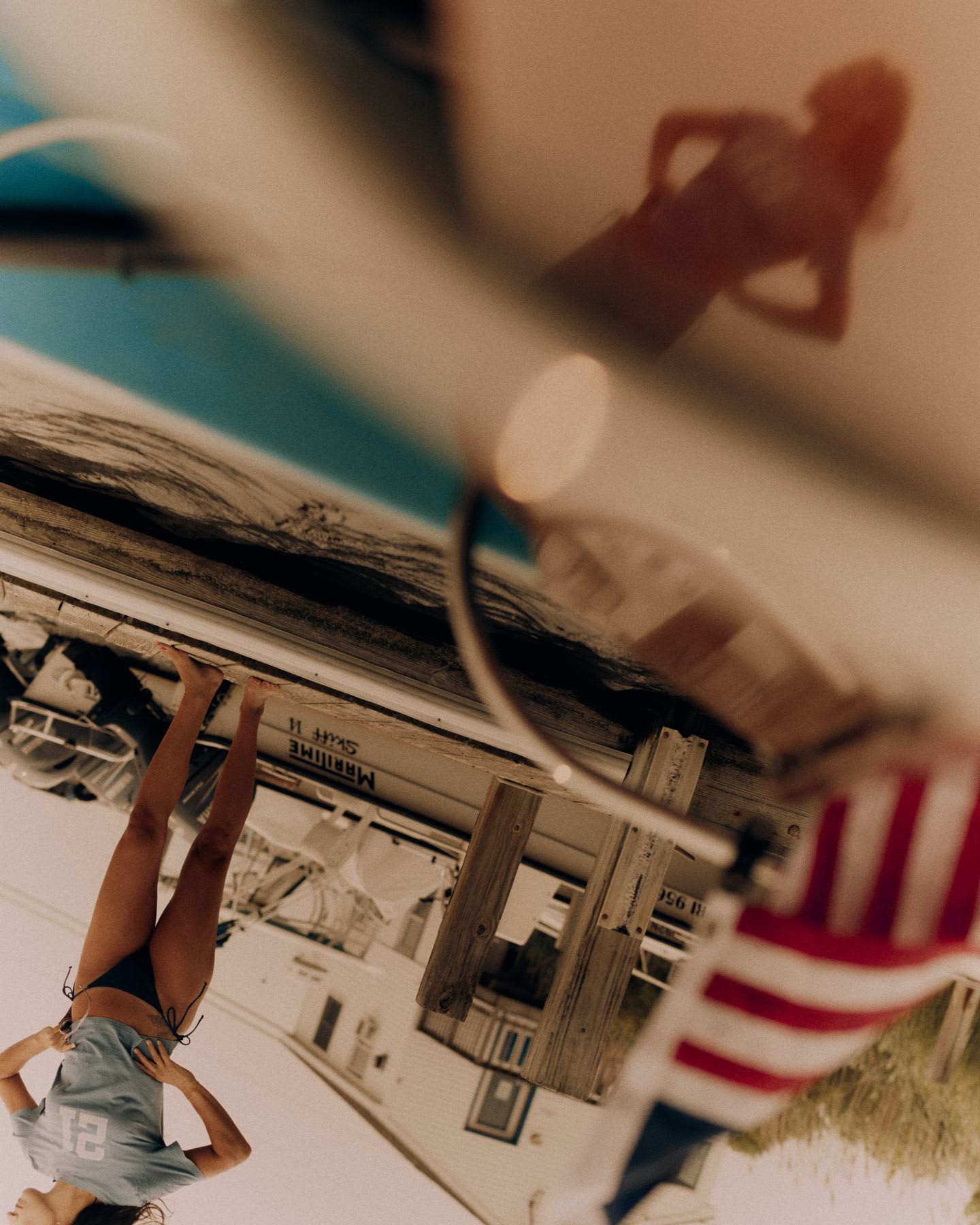 An artistic, upside-down photograph shows a woman in a t-shirt and bikini bottoms standing on a wooden dock, with her silhouette reflected on a shiny surface.