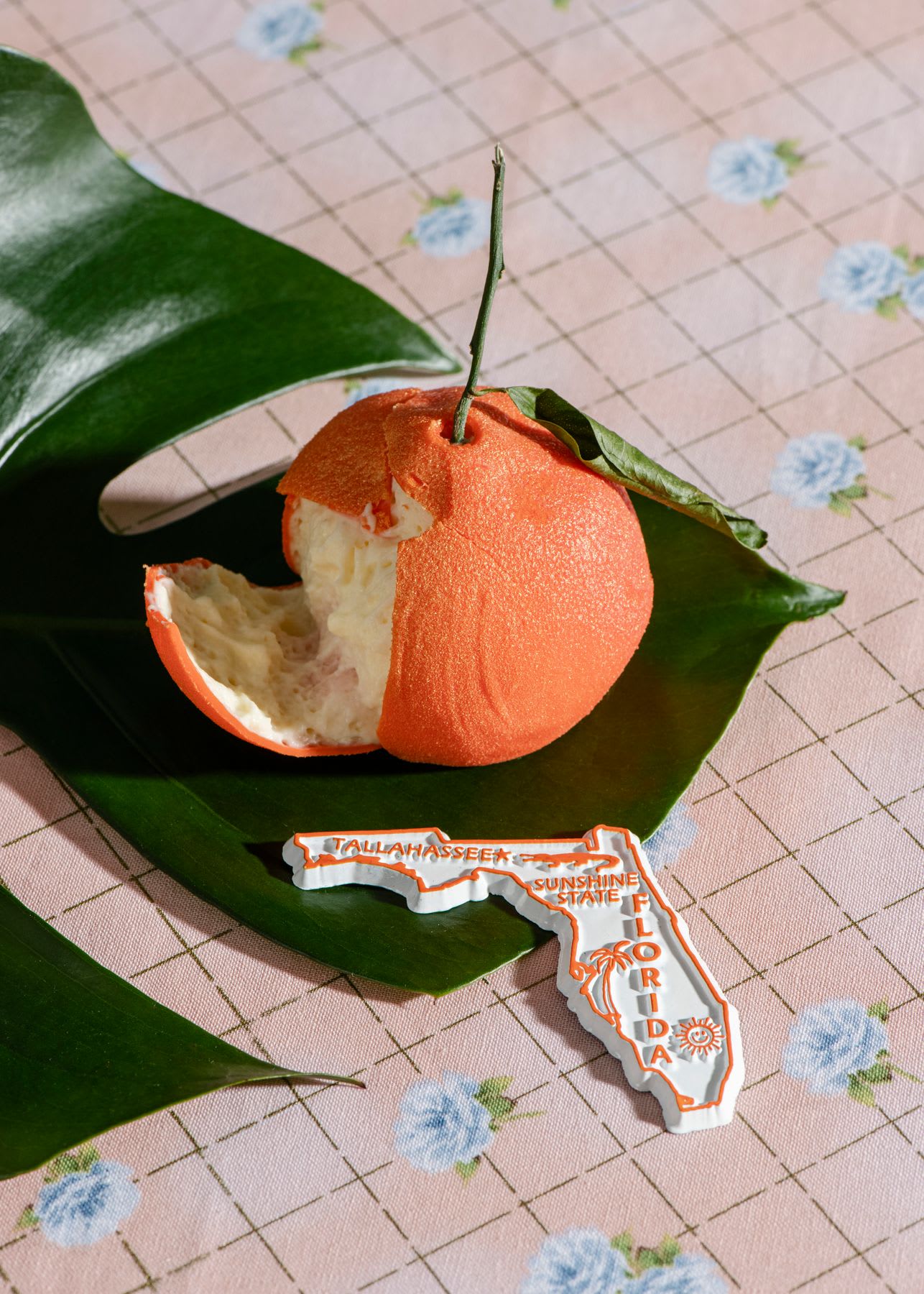 A still life composition of a partially peeled orange and a Florida state souvenir resting on a tropical leaf on a pink checkered tablecloth.