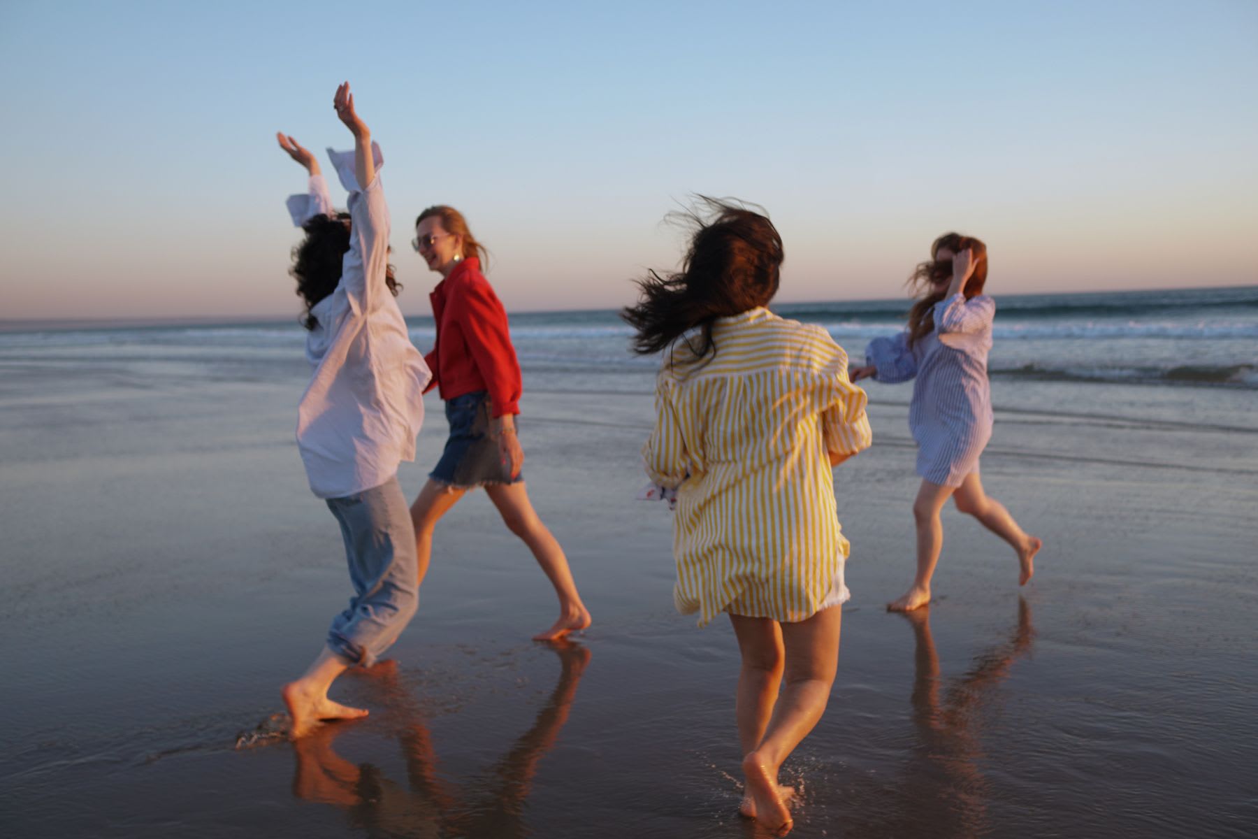 Four women enjoy themselves by the sea during sunset, with beach waves in the background.