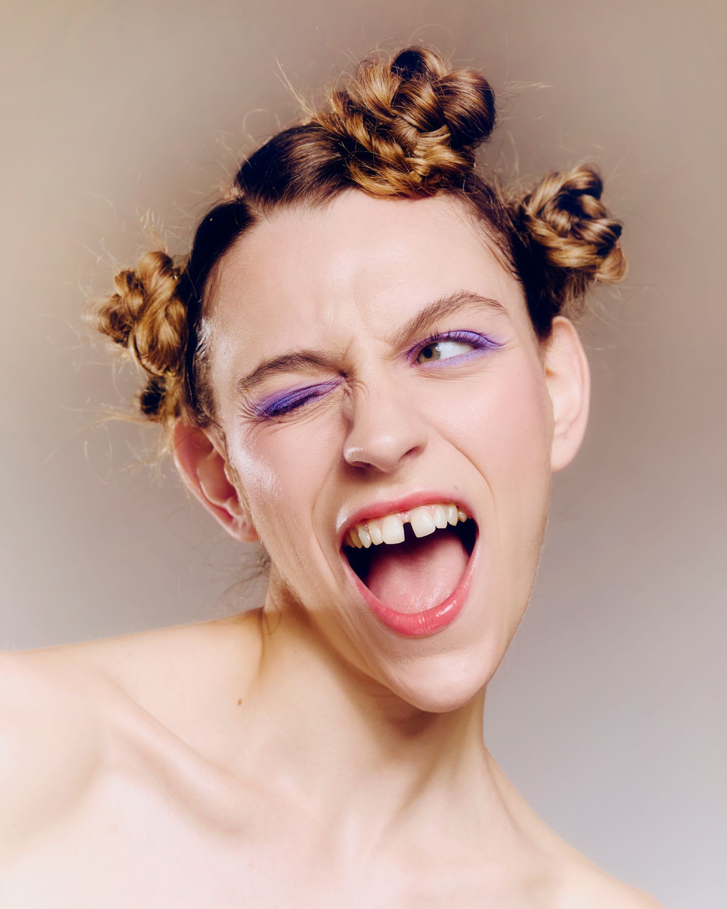 An expressive woman with braided space buns and purple eyeliner winks and shouts, revealing a gap between her teeth.