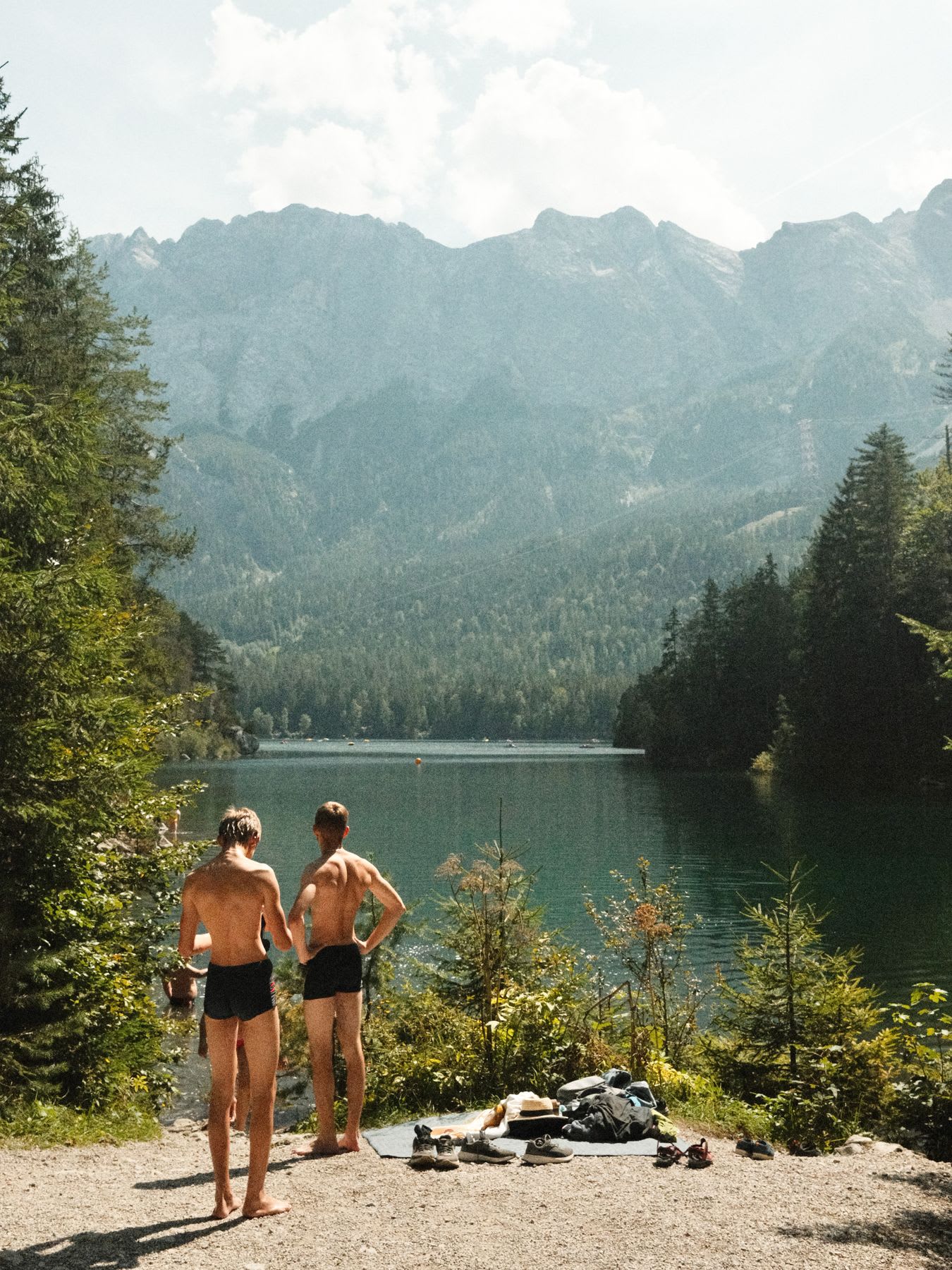 Two men in swim trunks stand on the shore of a beautiful mountain lake, preparing to swim on a sunny summer day.