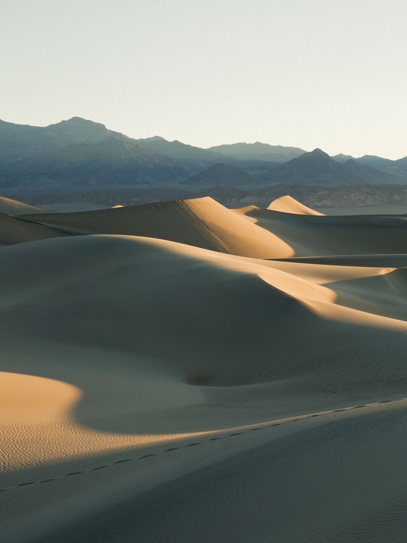 Desert sandscape with towering dunes glows as the sun dips below mountains, leaving a trail of footprints in its wake