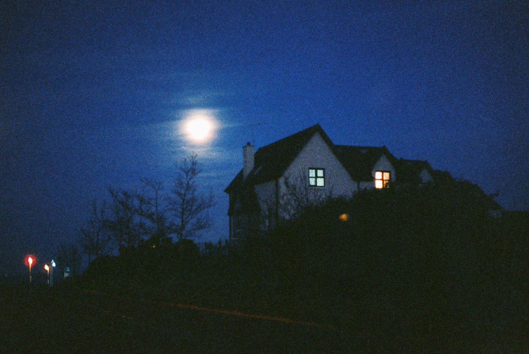 Under a bright full moon in a deep blue night sky, a silhouetted house sits atop a hill, with a single lit window glowing warmly and a few small lights twinkling in the distance.