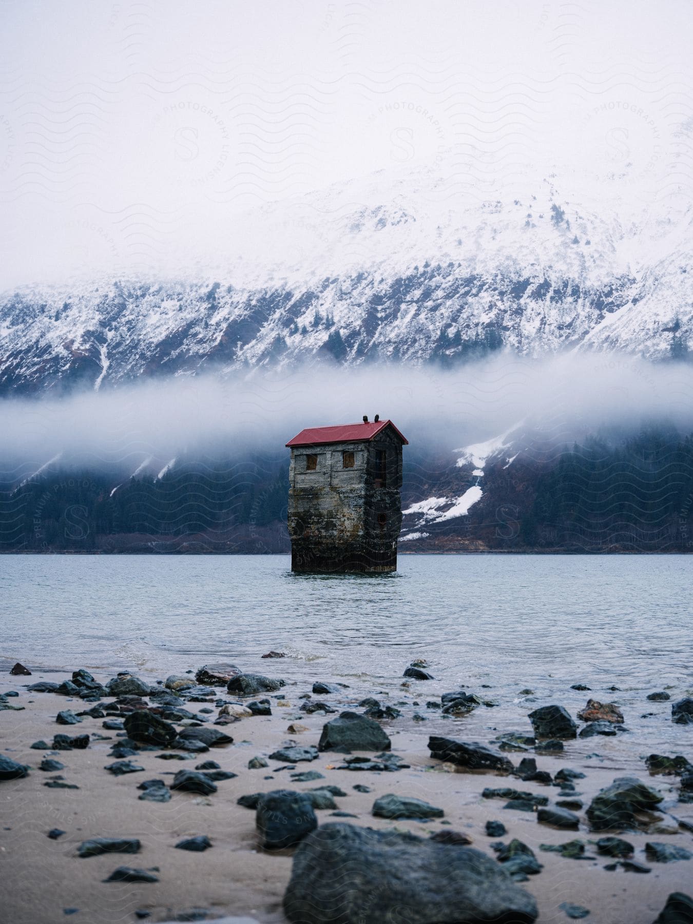 A tall, weathered cabin sits in the middle of a lake with snow-covered mountains in the background.