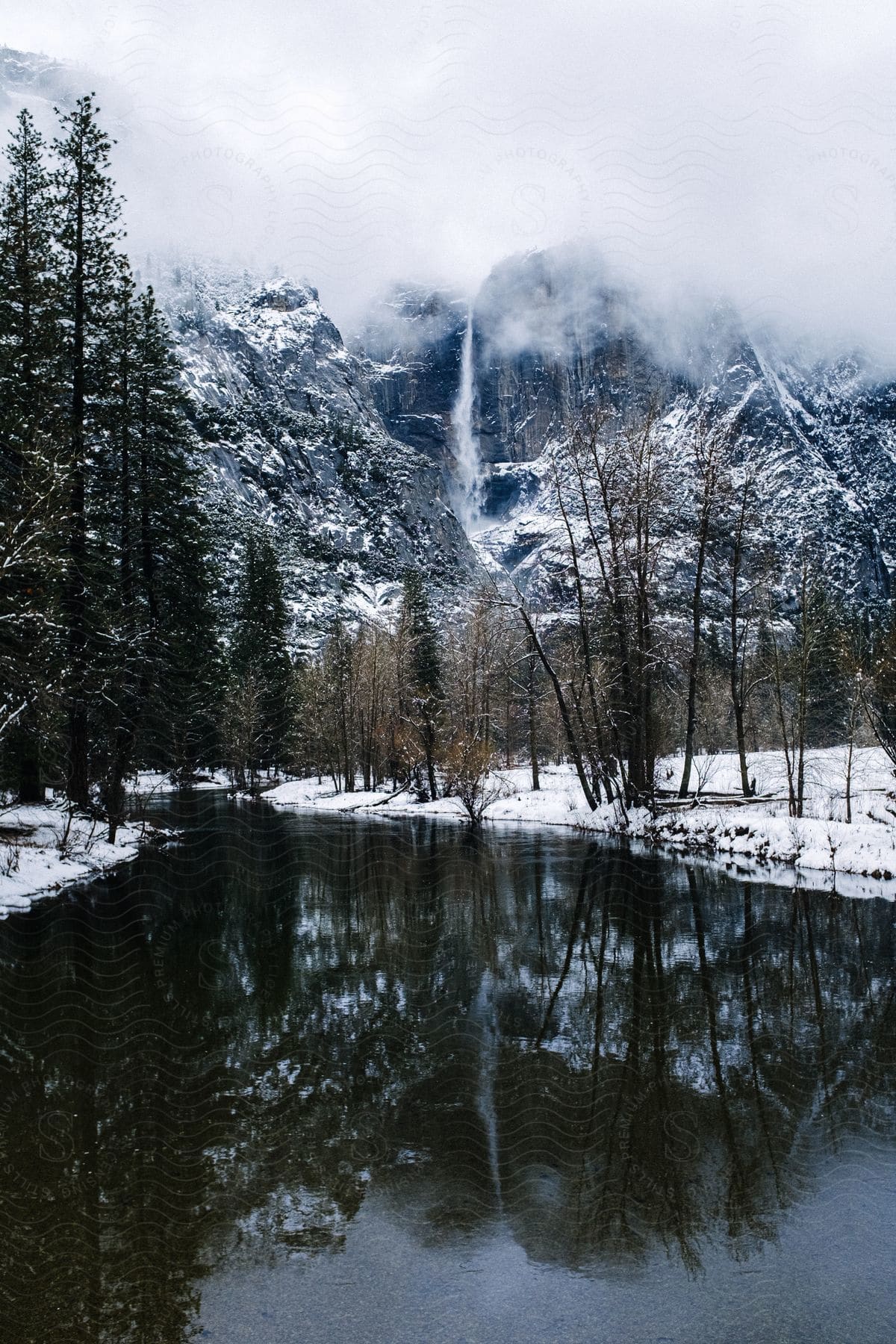 A tall waterfall flows over a mountainside during the winter with a still stream below.