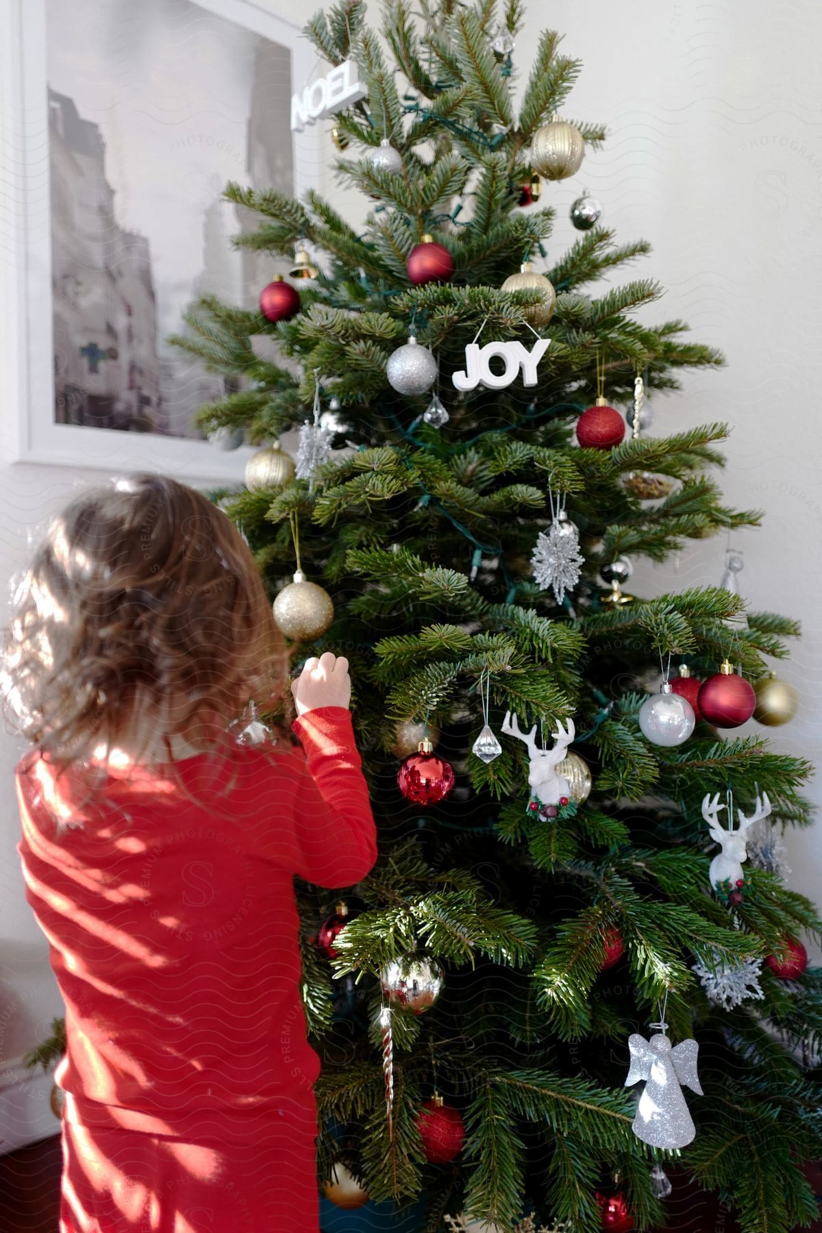 A girl with wavy hair, dressed in red, decorates a Christmas tree.