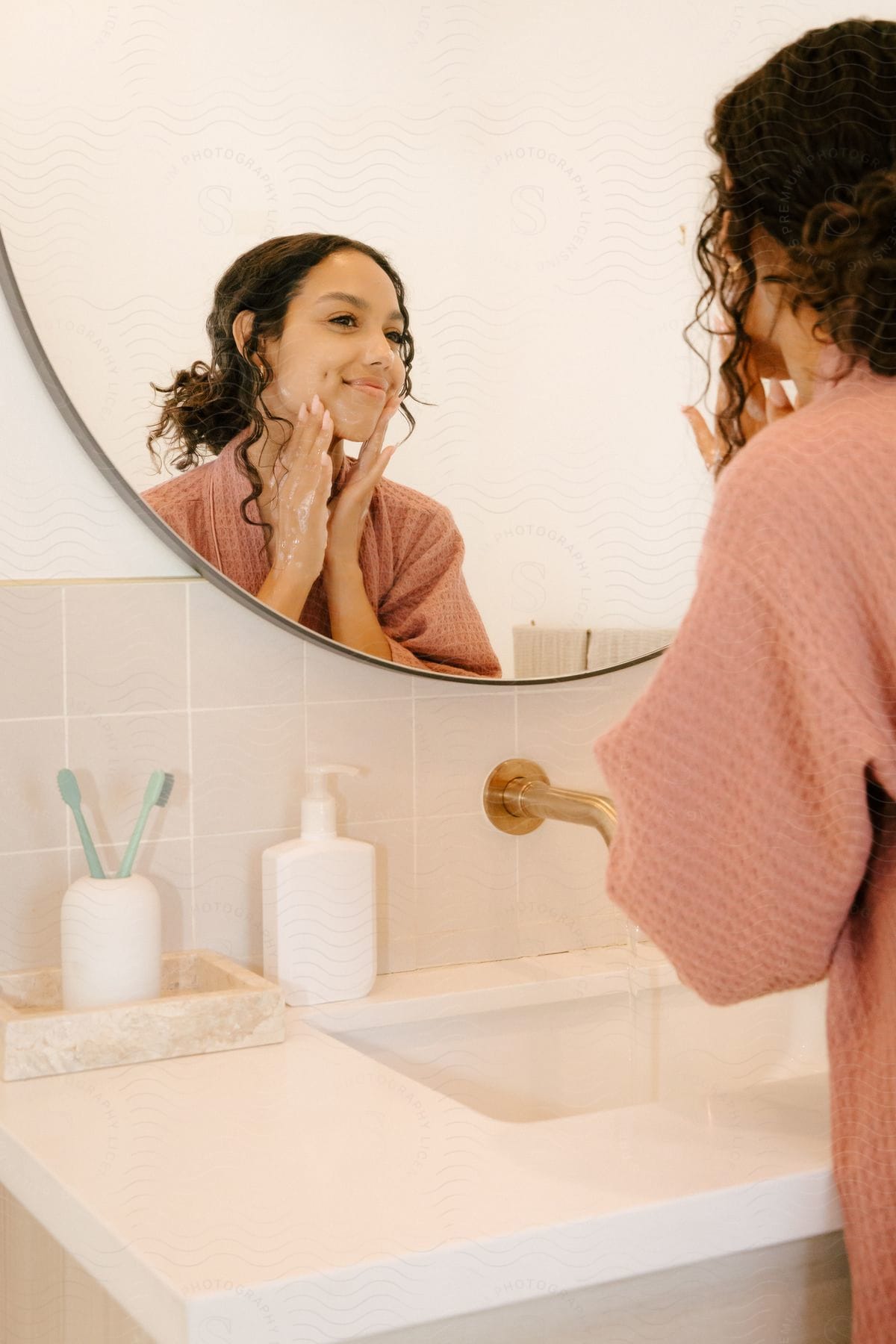 Woman washing her face with a smile in front of the mirror in the bathroom.