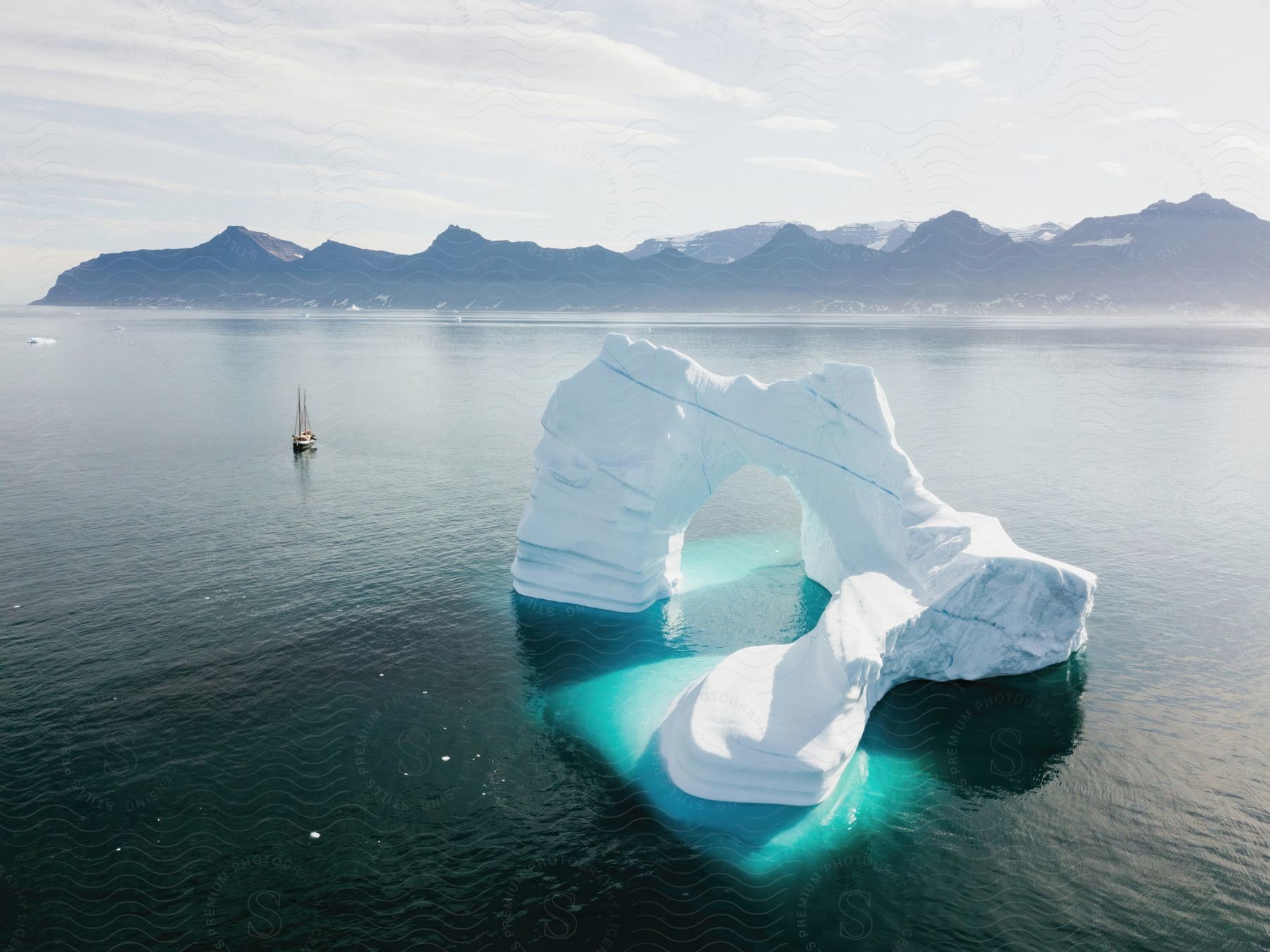 A sailboat travels next to an iceberg near the ocean coast with a mountain range in the distance.