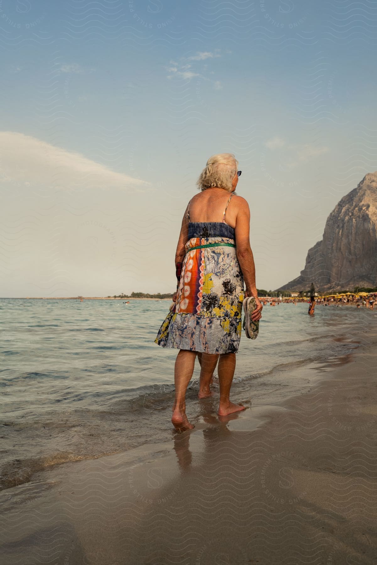 A woman walks on the beach as people are in the water near the coastal mountain