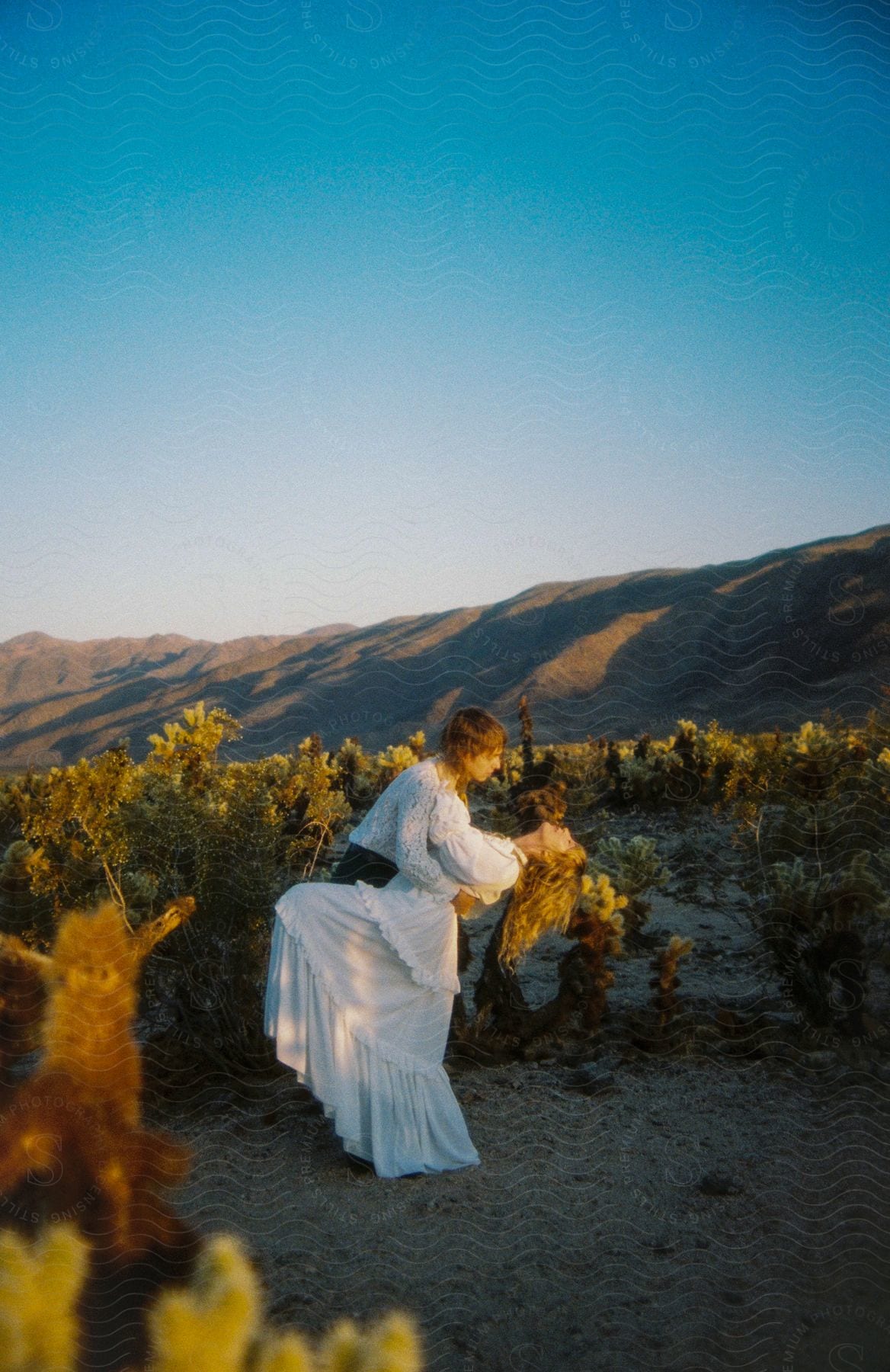 A young man holds a young woman in a white dress while surrounded by desert plant life.