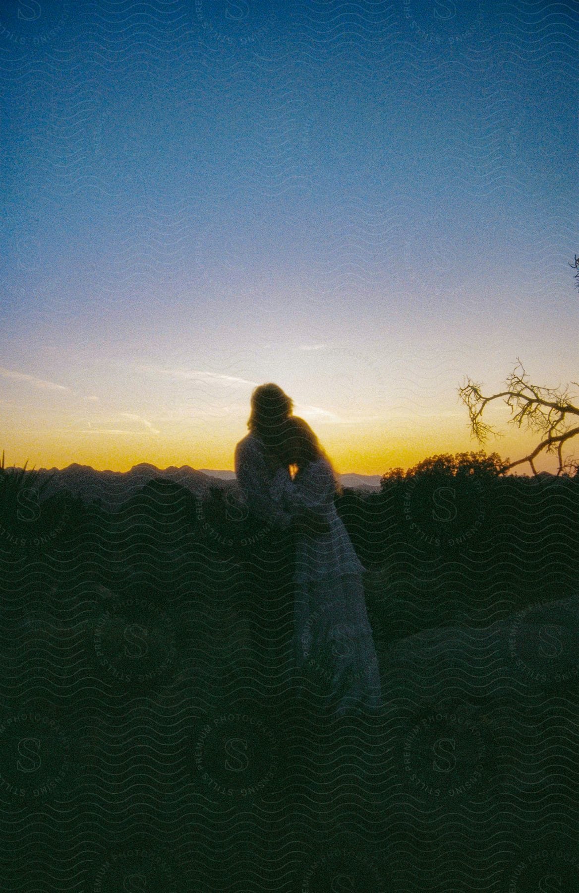 A man and woman are embracing as they stand on high ground and the sun sets with mountains in the distance