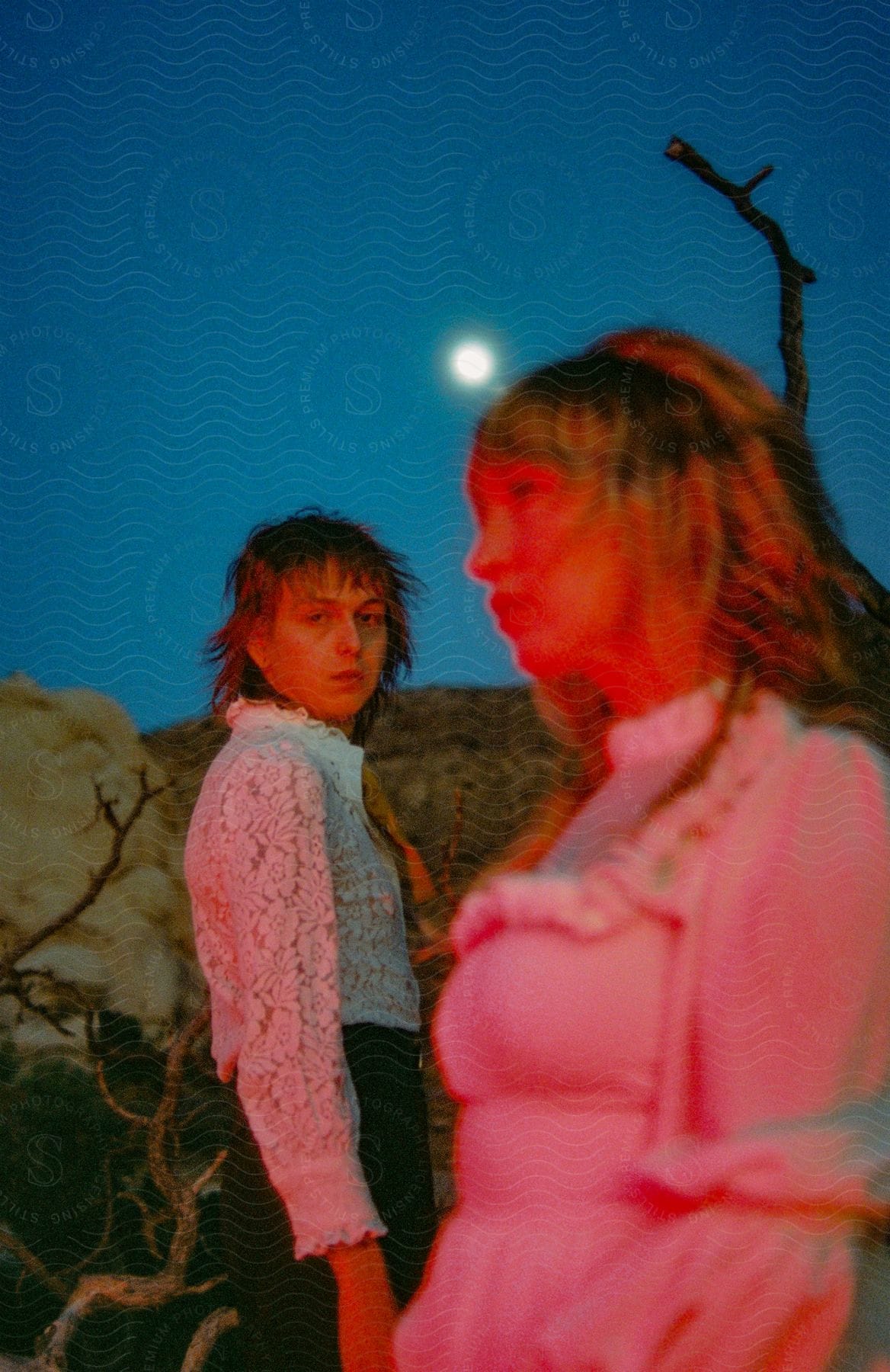 Two women, one in a white dress and the other in a blouse, stand beneath the shining moon in the desert.