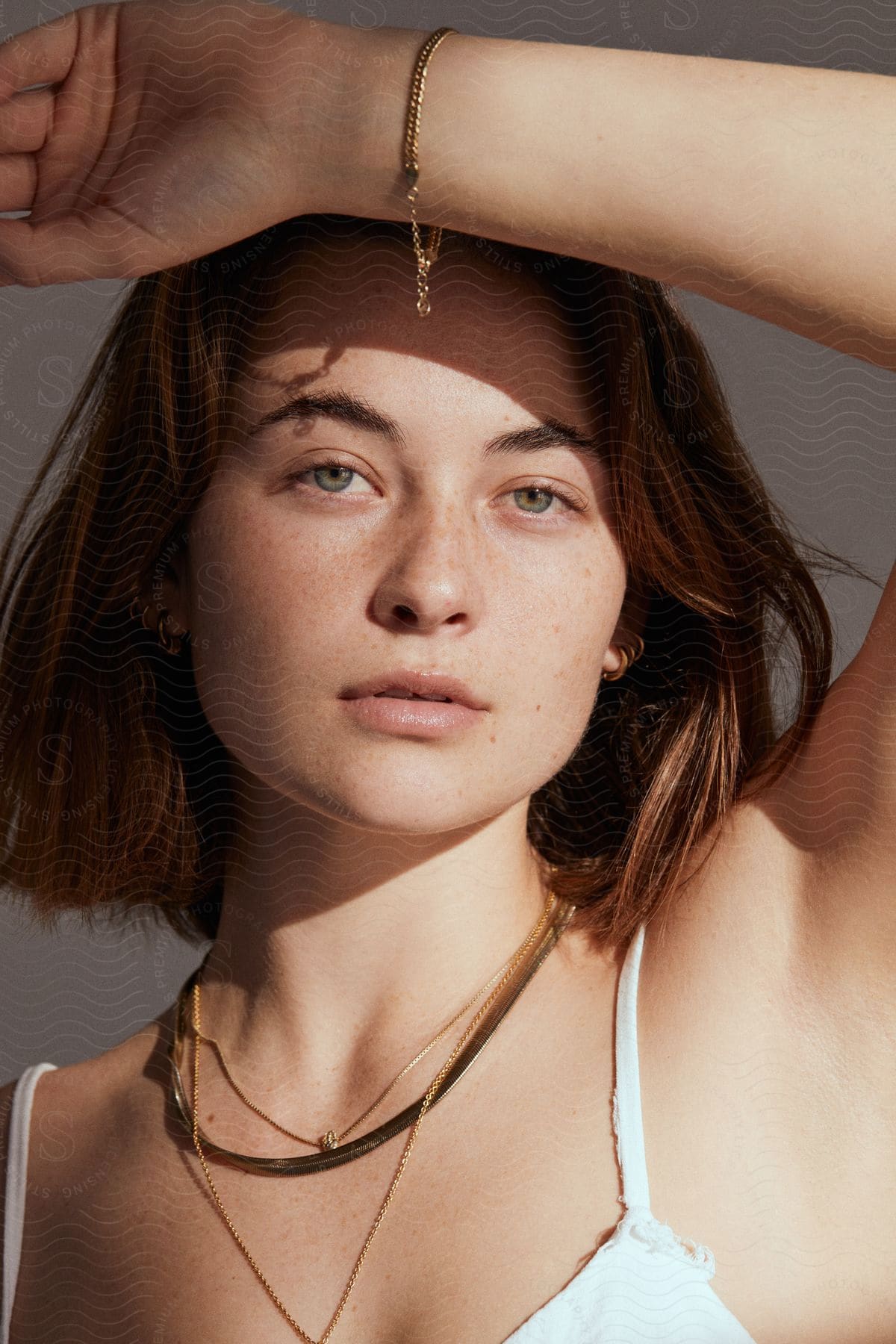 A woman with brown hair, wearing pendants and a white top, rests her wrist on her forearm