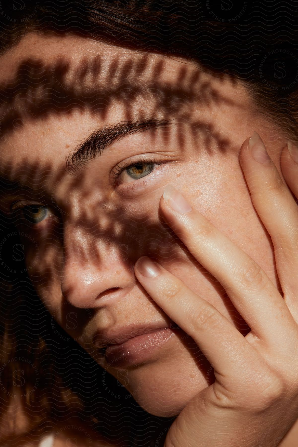 Portrait of a light-eyed woman with hands on her face, contemplative, and with tree leaf shadows on her face.