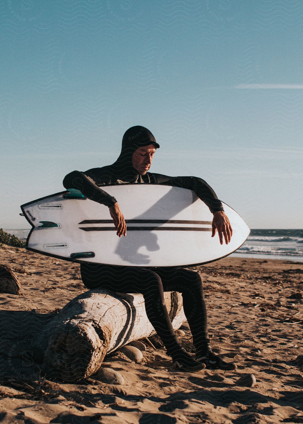 Man in a wetsuit sitting on a wooden log on the beach, holding his surfboard.