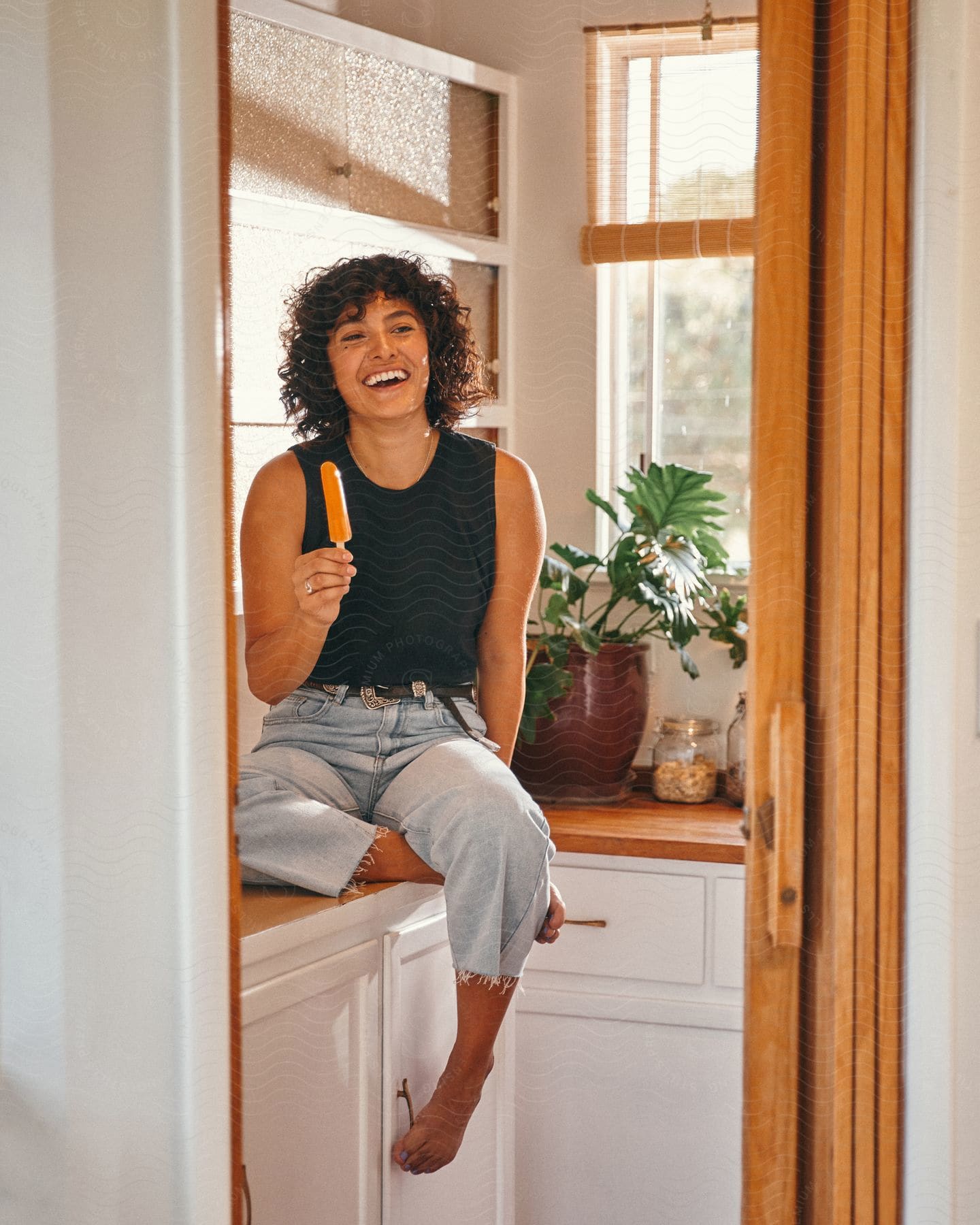 A woman is sitting on the kitchen counter laughing as she holds a popsicle