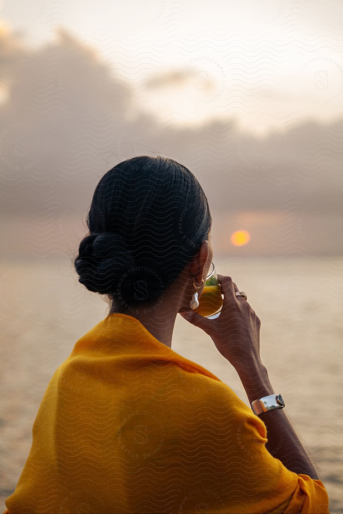 A woman in yellow clothing is drinking something while looking at the sea horizon with the sun above.