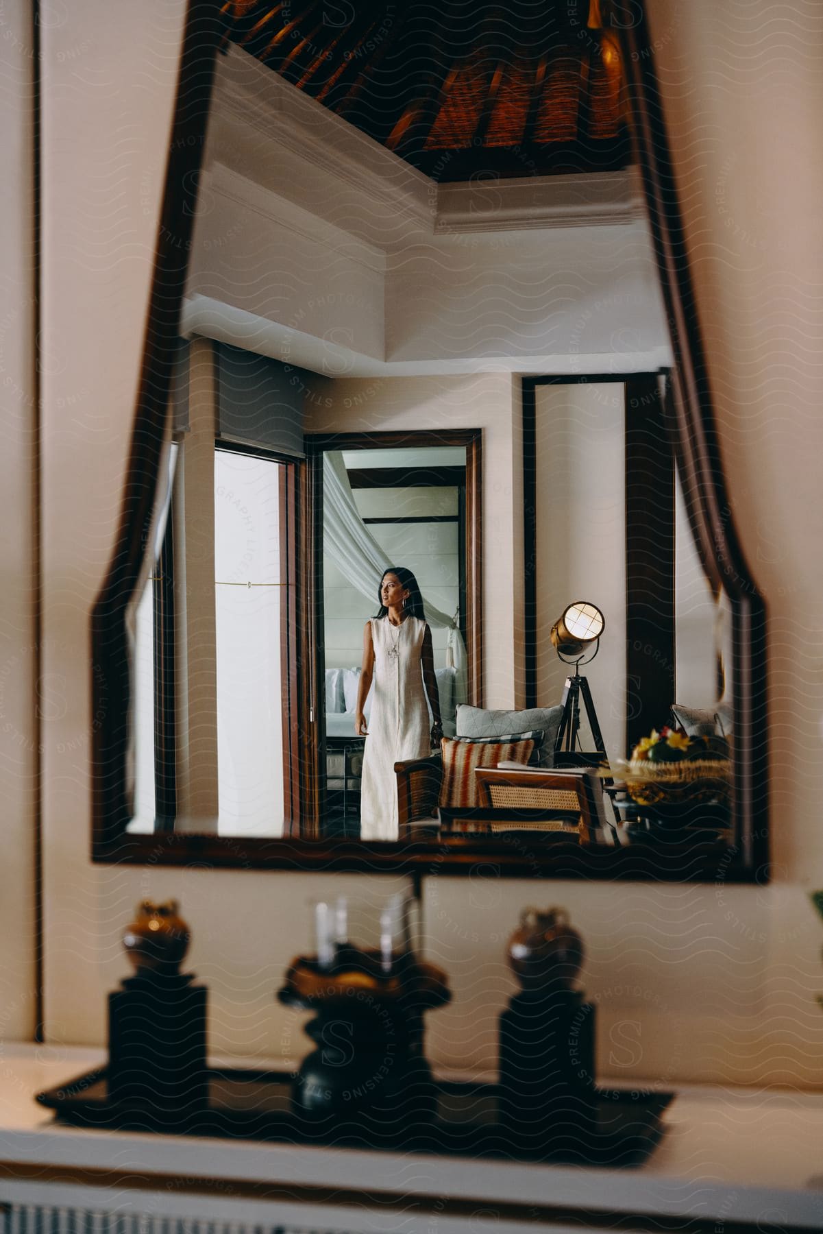 A woman wearing a white dress is seen in a mirror standing in the doorway of a room