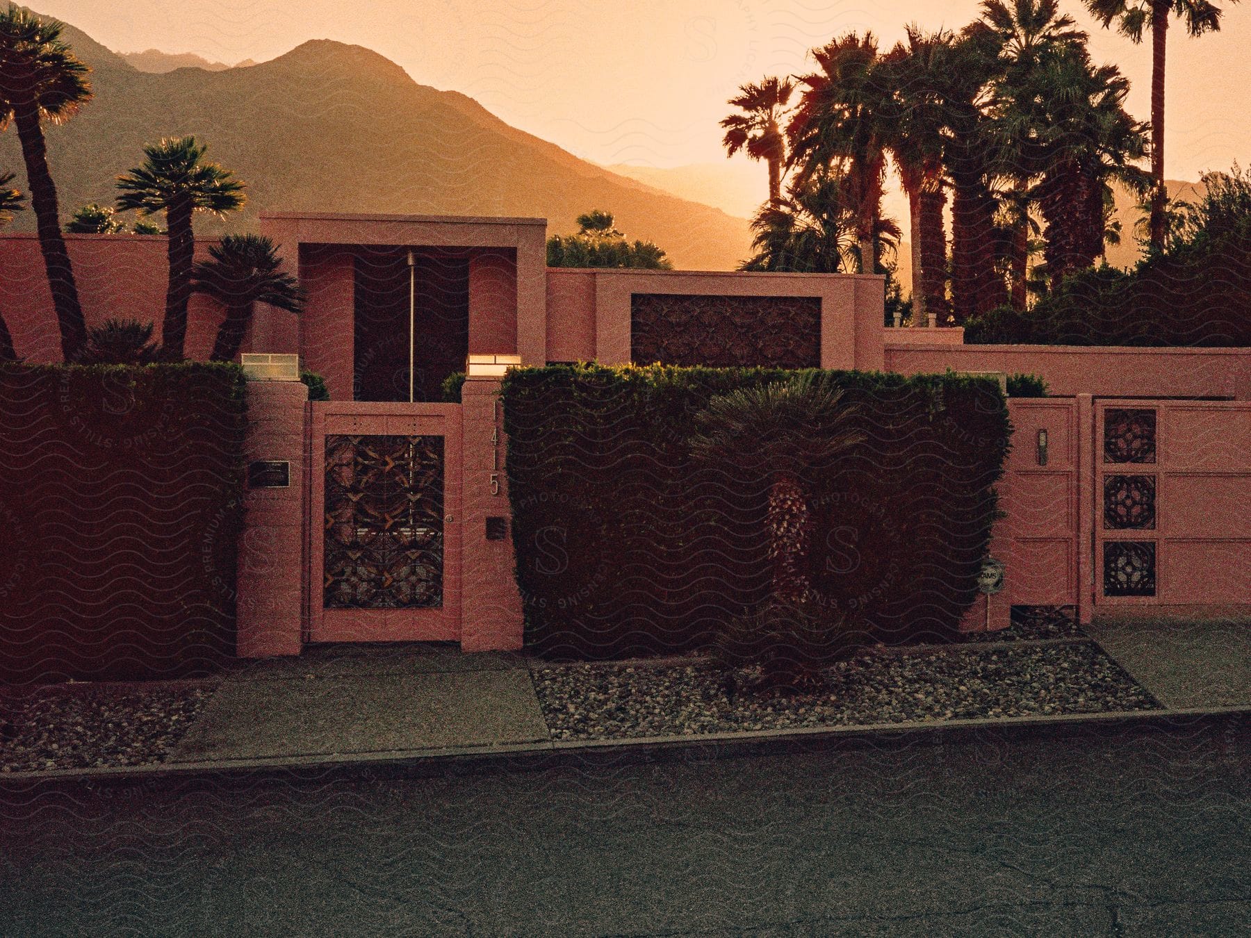 A house featuring a pink brick wall, flanked by palm trees, set against the backdrop of a mountain range