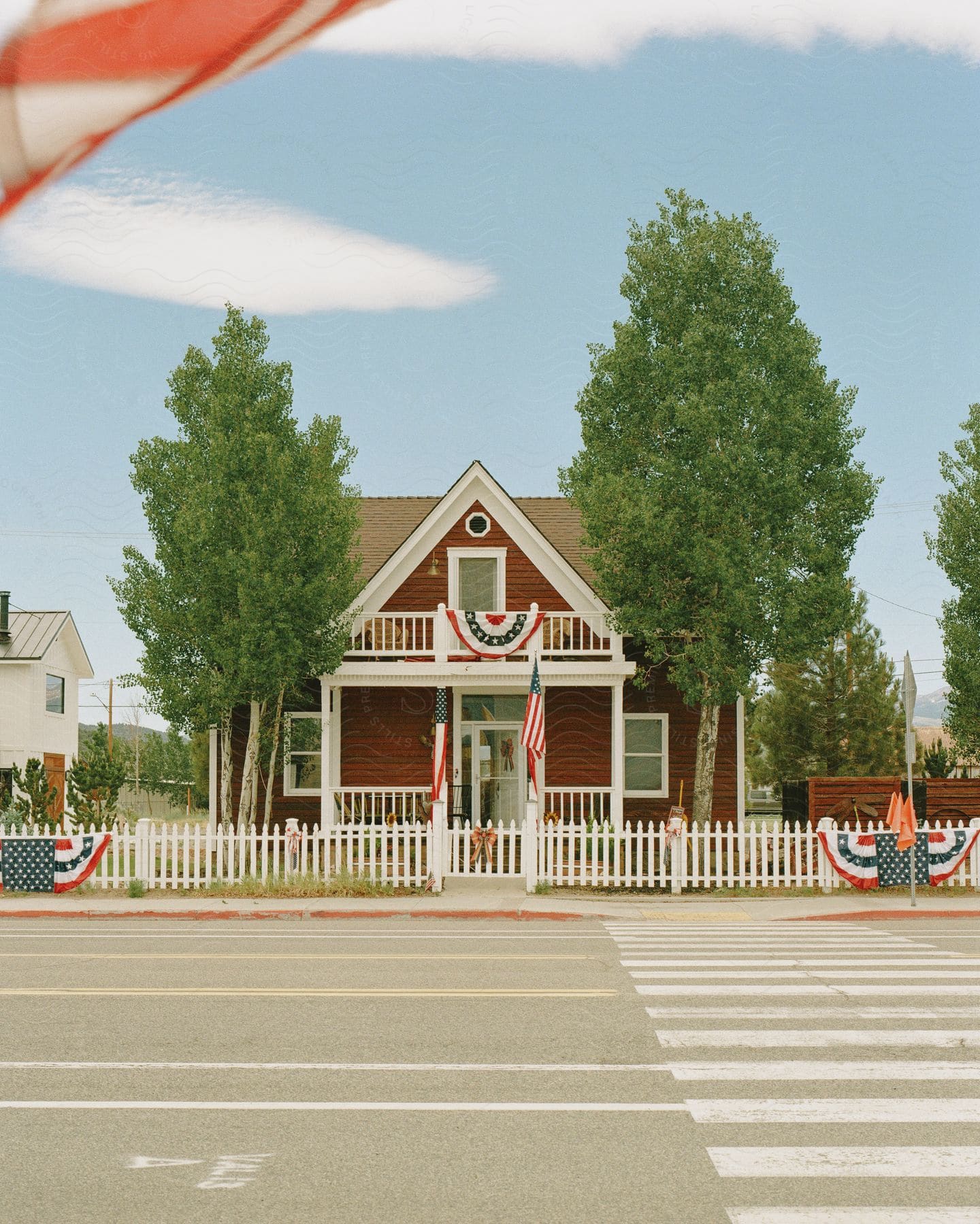 A house is behind a white picket fence with two trees and flying American flags