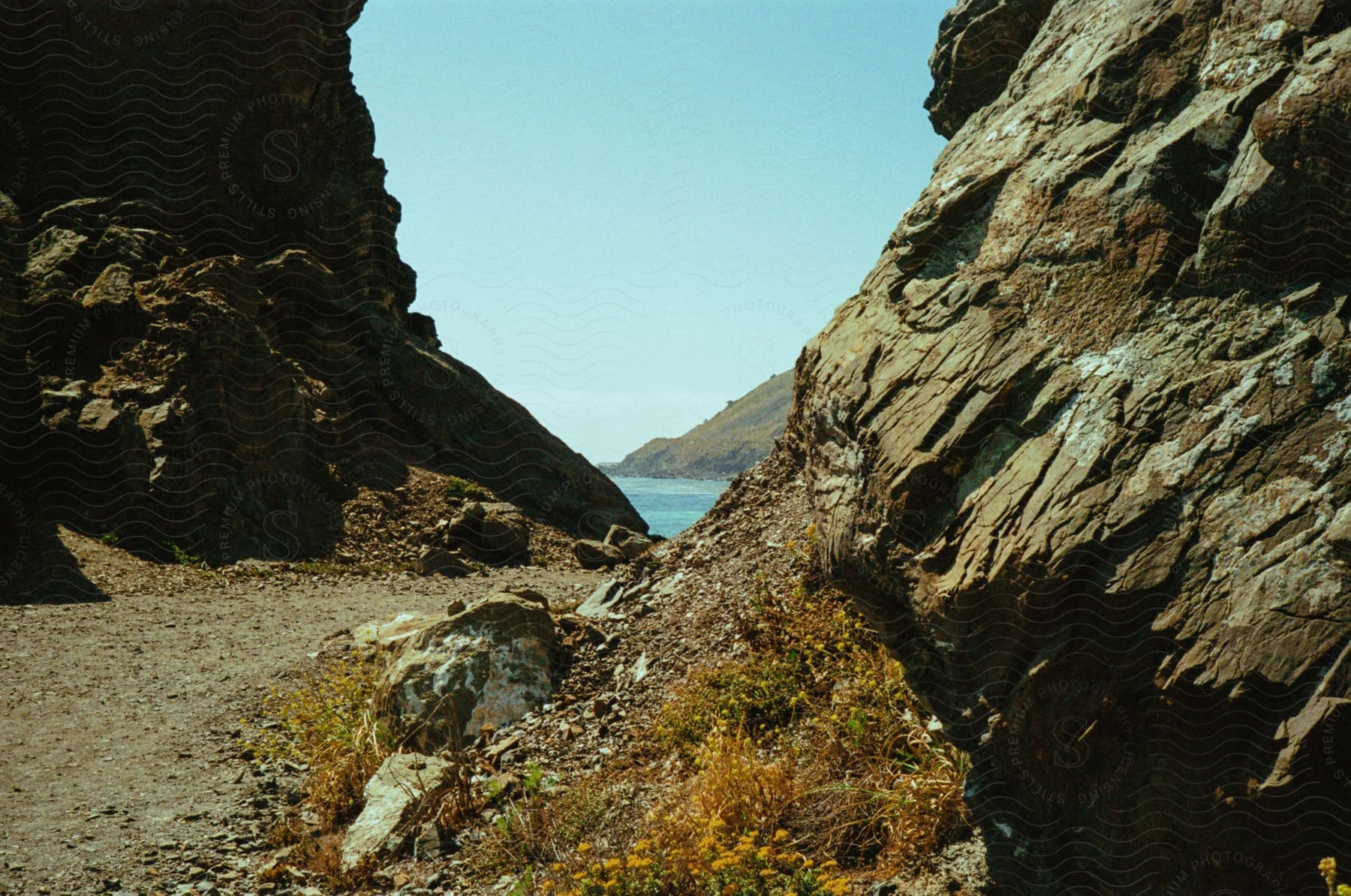 A dirt path surrounded by mountain rocks and a blue sea in the background.