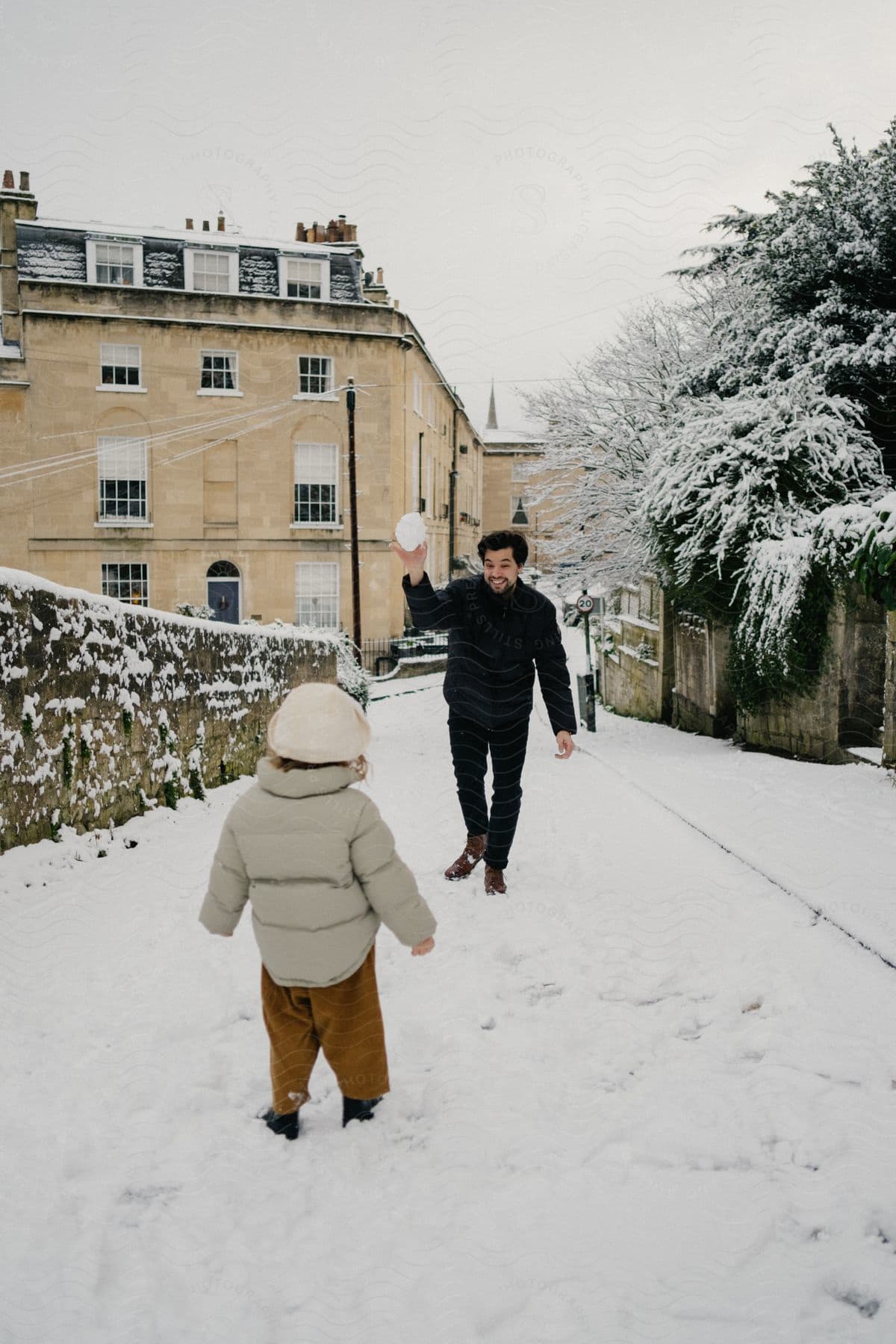 child stands on a snow covered neighborhood street as a man holds up a big snowball with a smile on his face