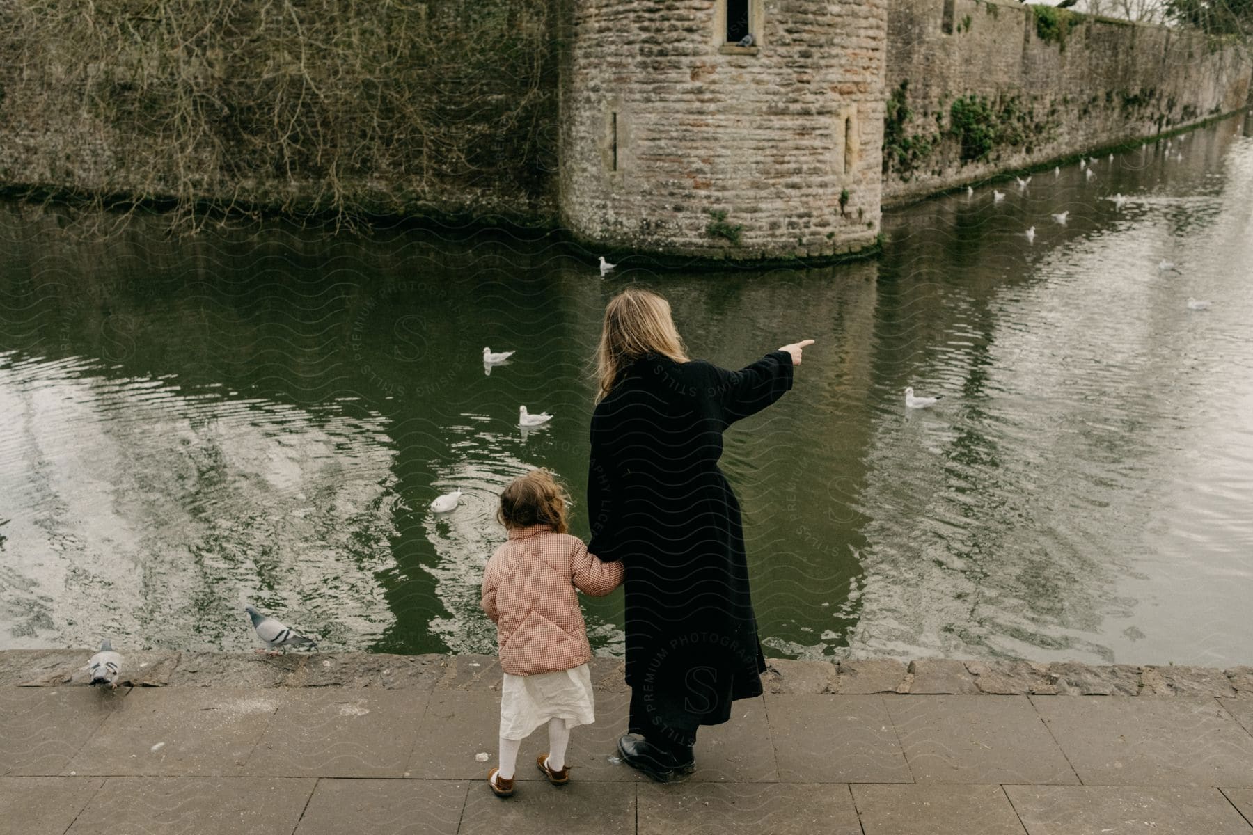 A woman is standing near the canal holding her young daughters hand as she points to ducks in the water