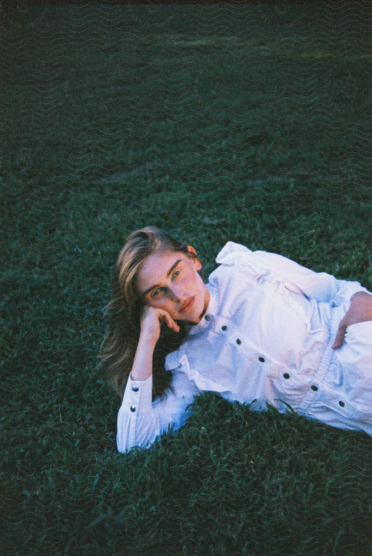 Blonde woman modeling in a white outfit, lying on the grass and resting her head on her hands.