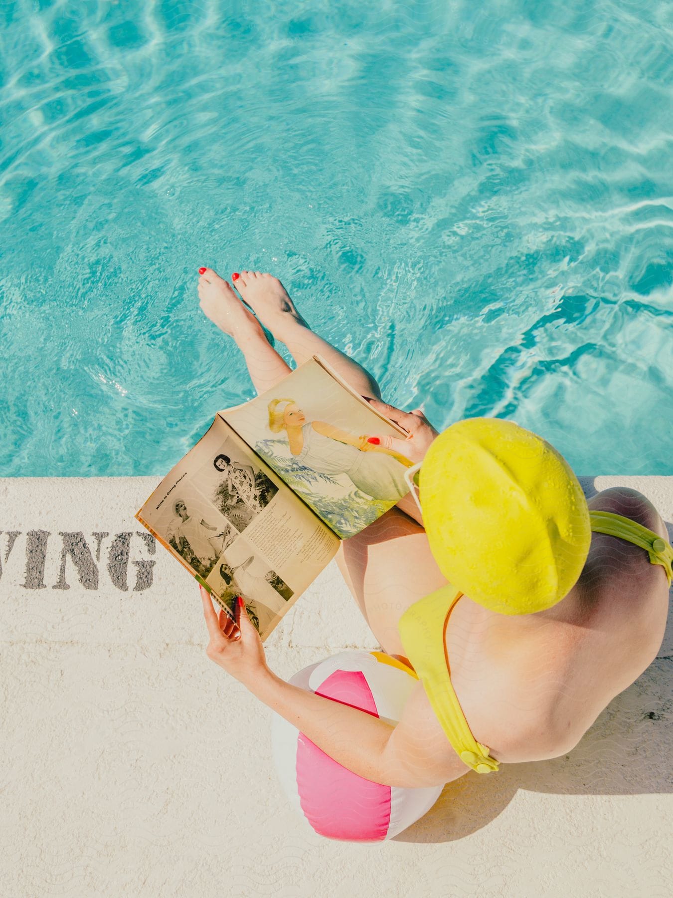 Woman in a swimsuit and yellow cap, sitting by the pool and reading a magazine.