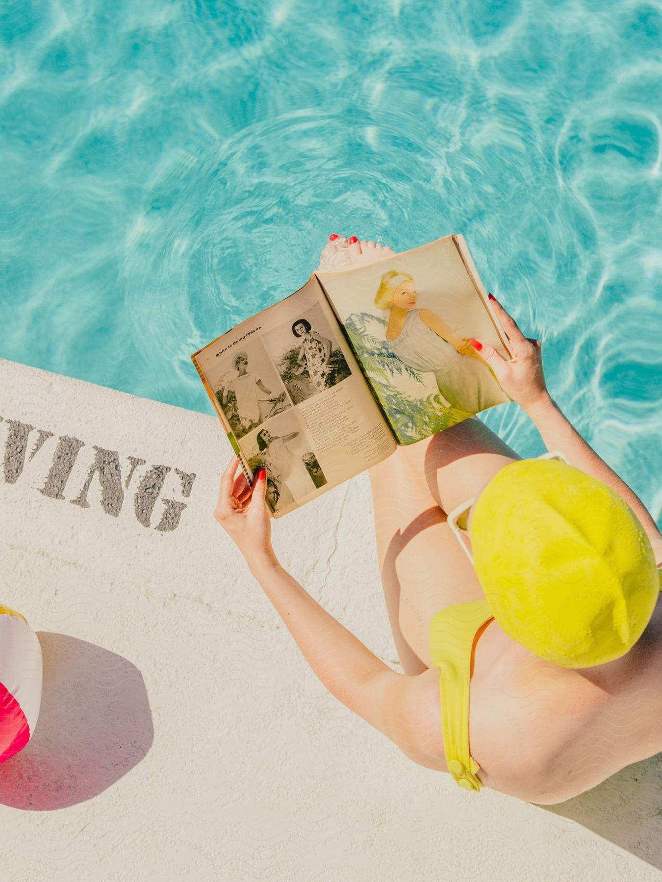 a woman wearing a yellow swimsuit sitting beside a pool as she read a modeling magazine