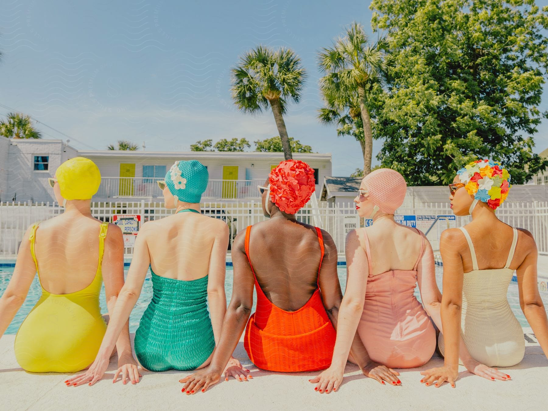 Five models wear bright-colored swimsuits while sitting next to each other poolside.