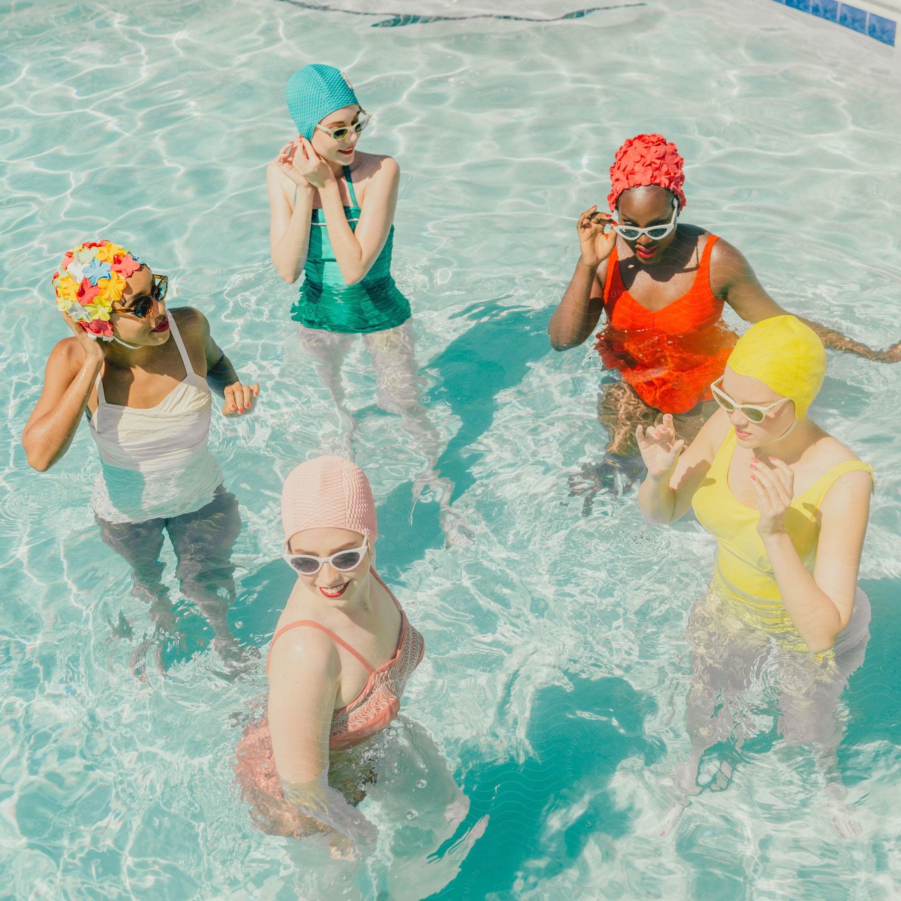 Five women in swimwear and colorful caps in a pool with crystal clear water.