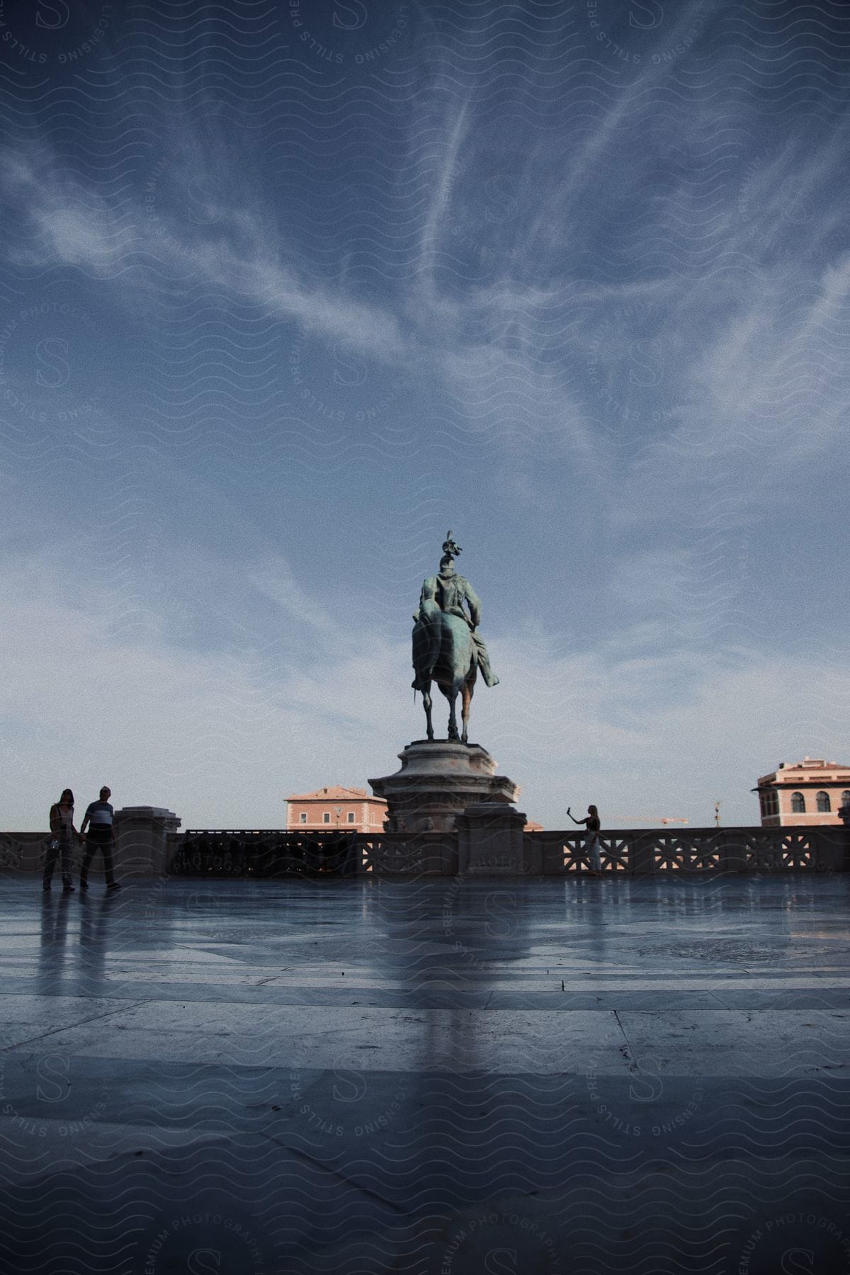 a monument status of a man on a horse as some couple walk by it and a lady by the status taking a photo during sundown