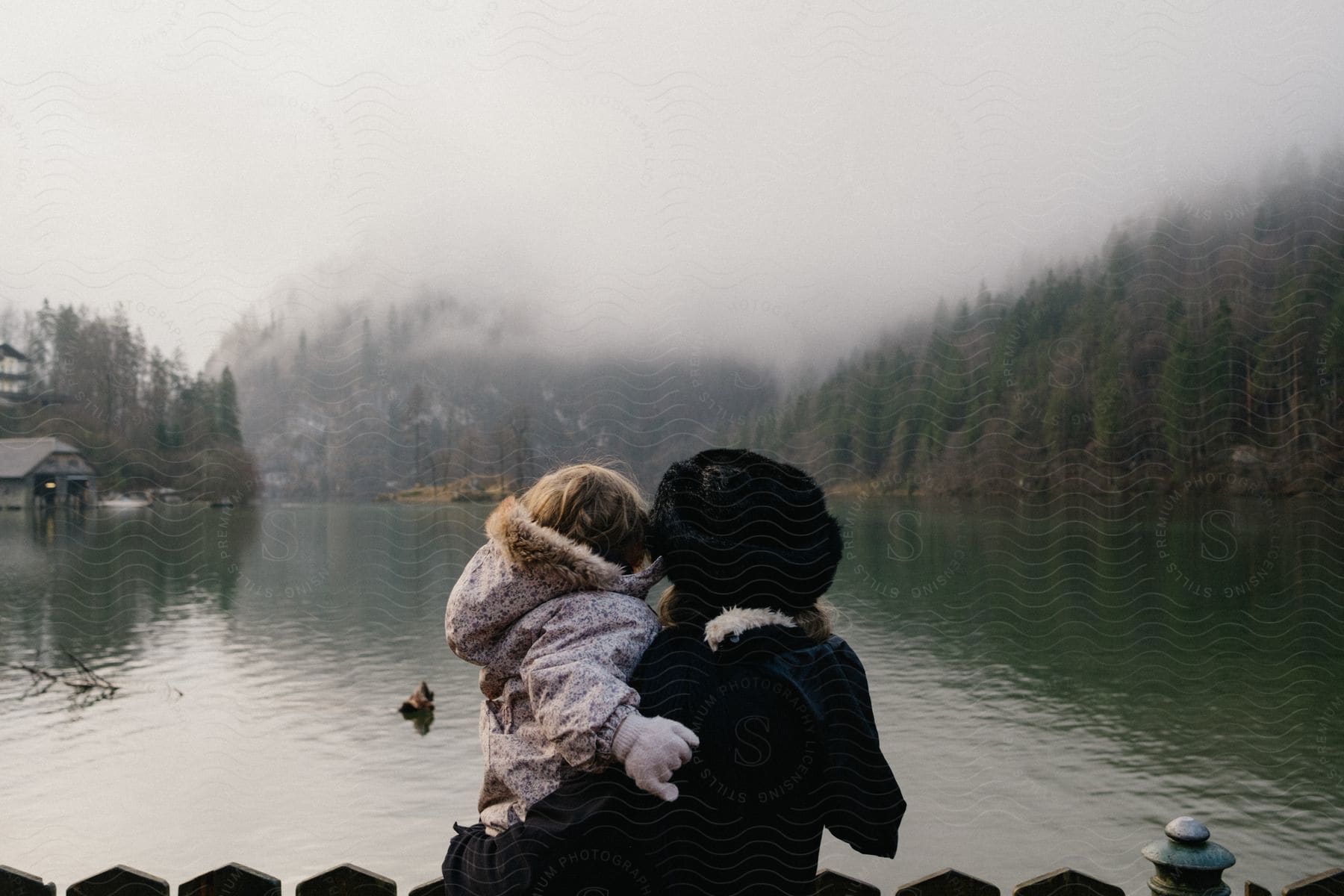 Mother holding her daughter in her arms, facing a lake with mountains and conifers in the background.