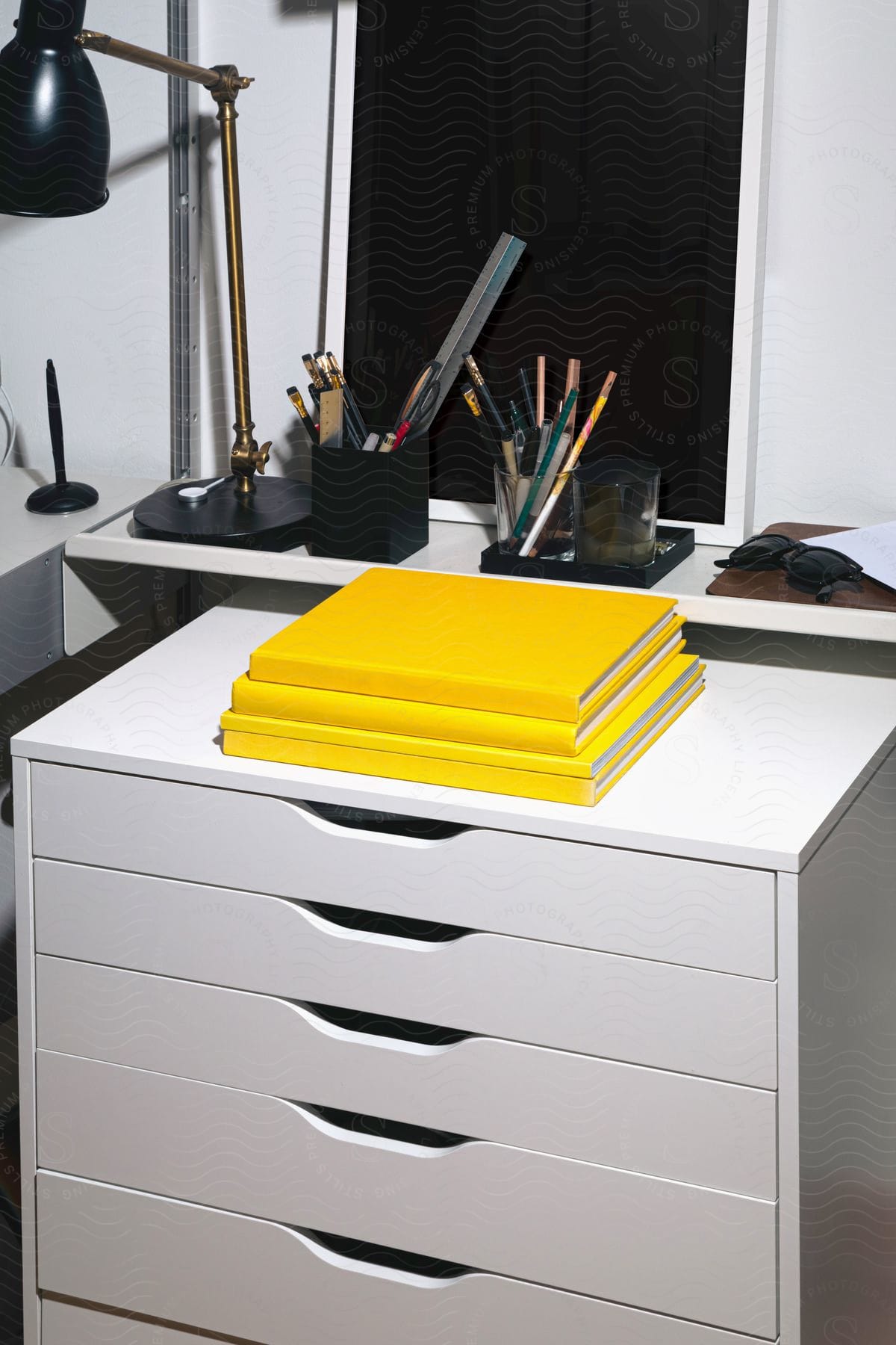 A White Desk With Yellow Books, A Table Lamp, And Containers With Pencils, Pens, Rulers, And Sunglasses Sits In Front Of A Blackboard