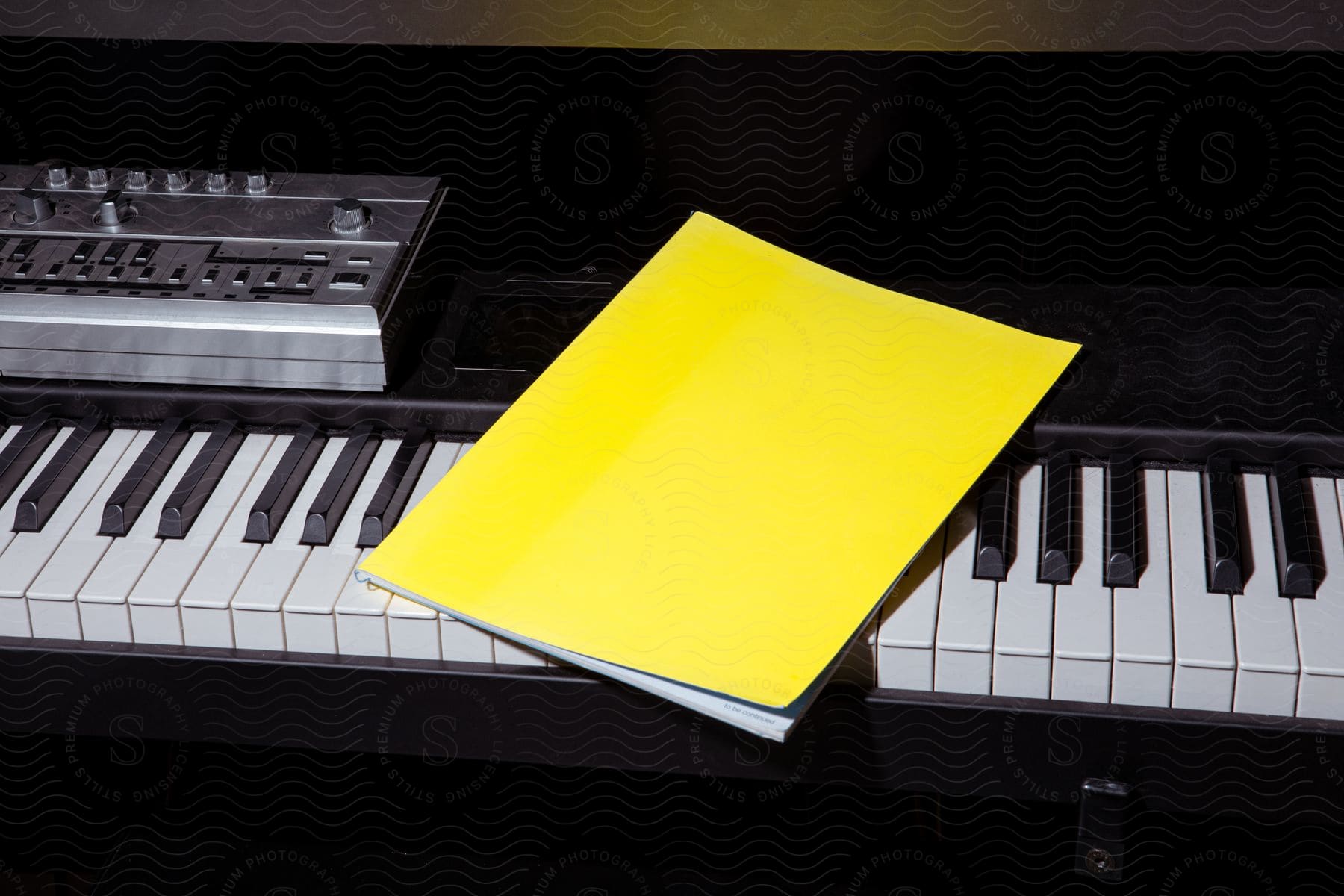 A bright yellow booklet is laying atop an electric piano in a darkened room.