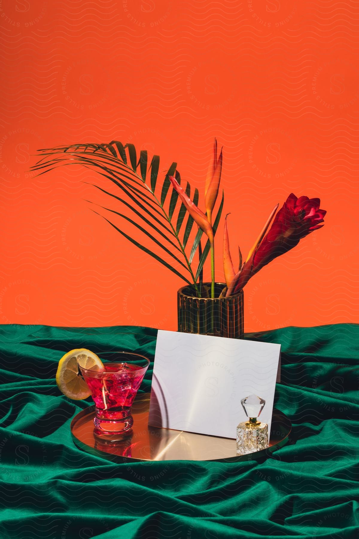 A pink cocktail sits on a tray with a bottle of perfume and a letter. Behind is a vase with flowers.