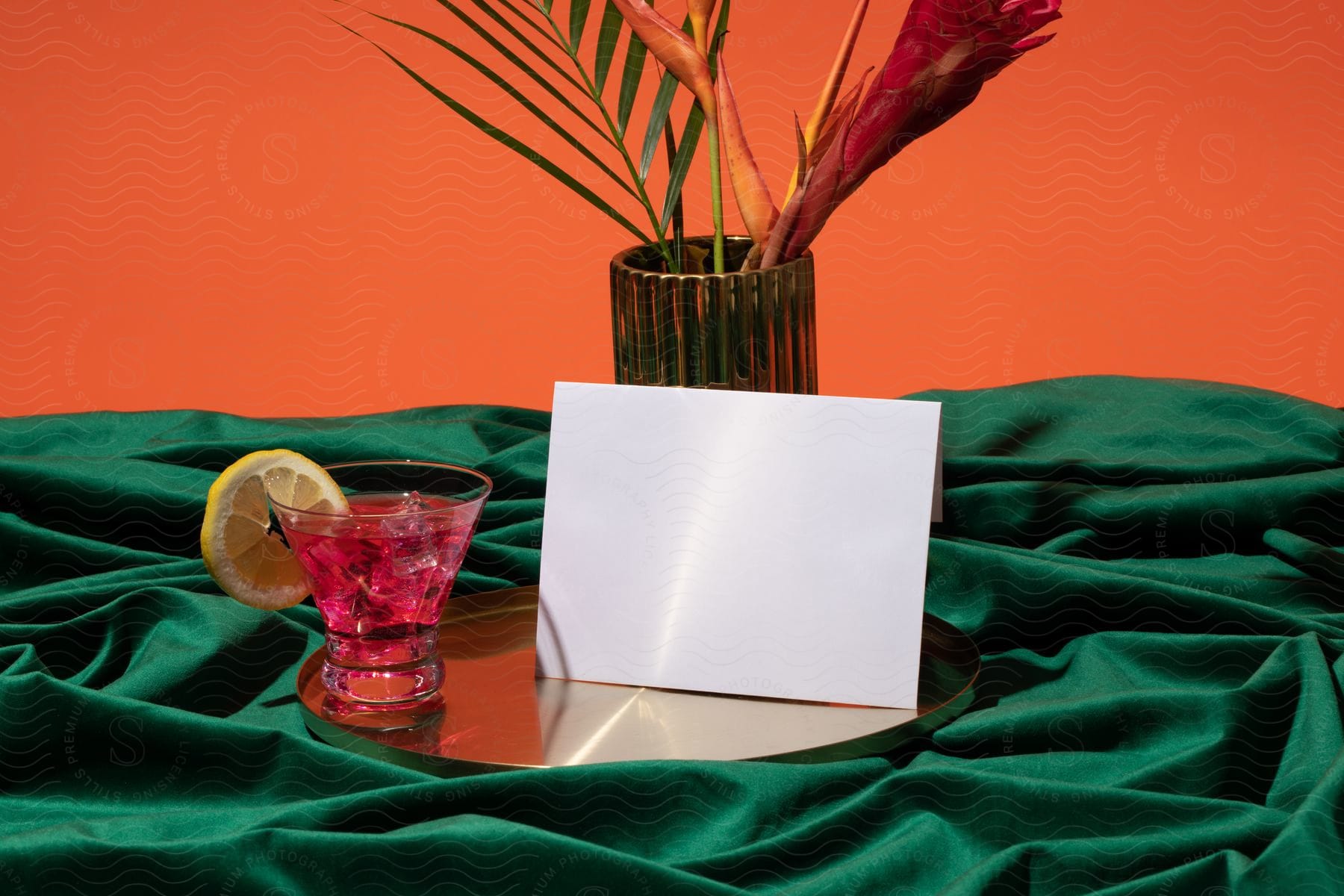 A single cocktail in an ice-filled glass sits on a tray with a plant and blank envelope.