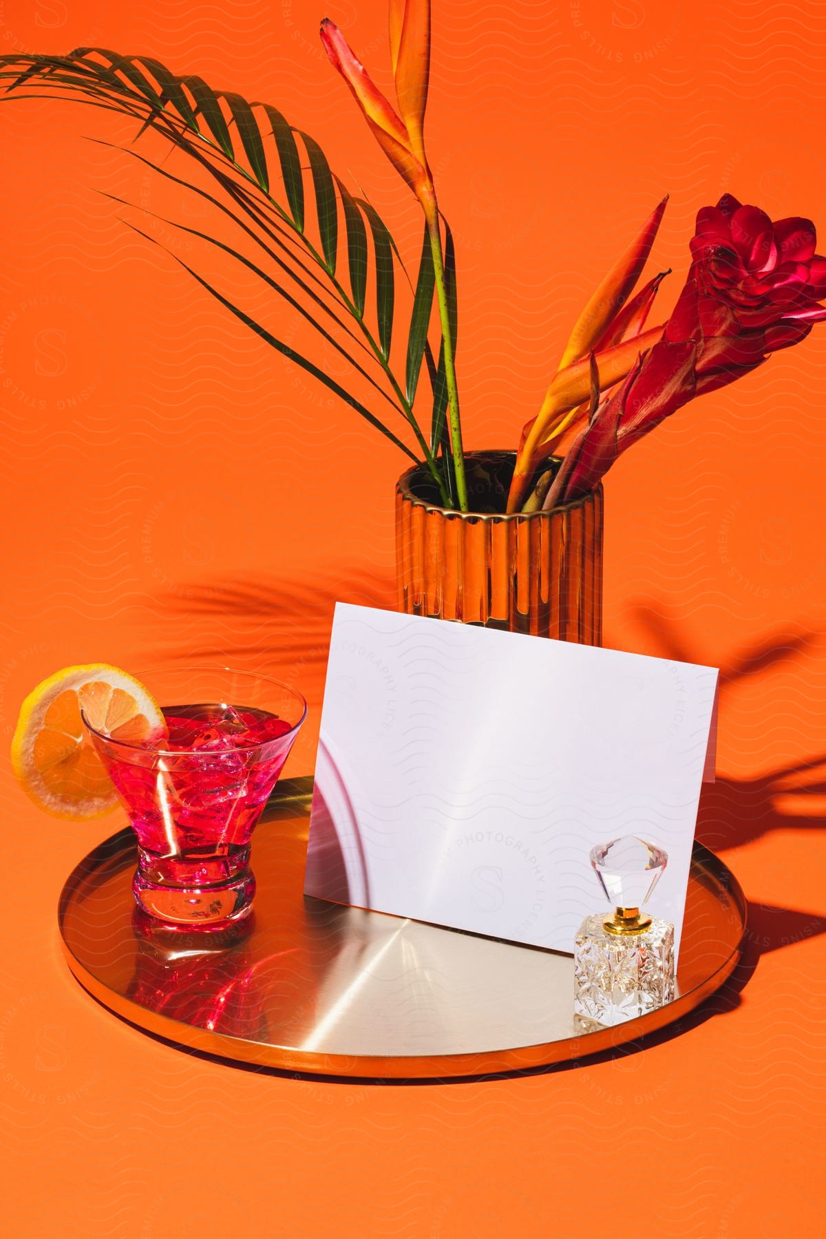 Stock photo of a cocktail served on a glass with a slice of lime and place on a tray next to an envelop and ring case with a flower behind on an orange background