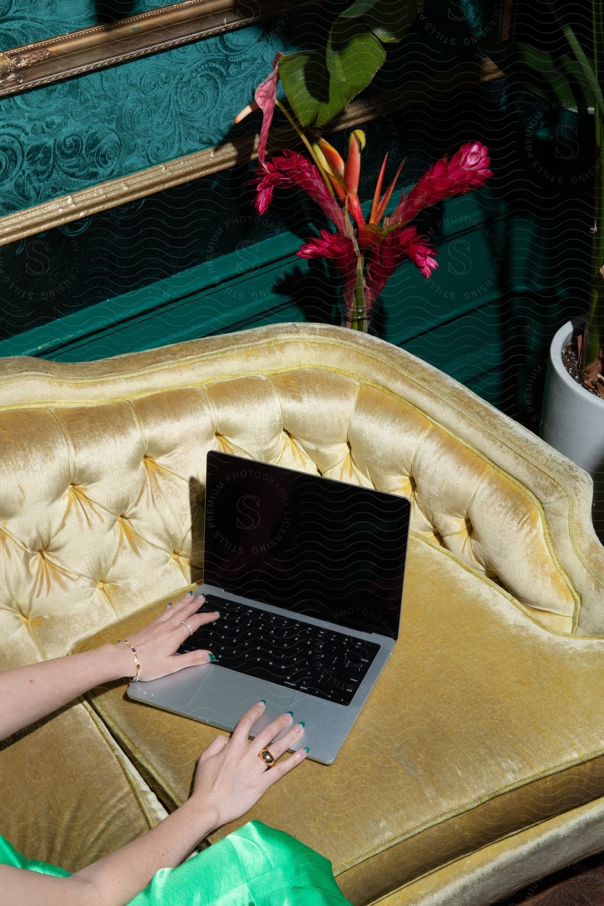 Stock photo of woman wearing a green dress uses silver laptop on yellow sofa near houseplants