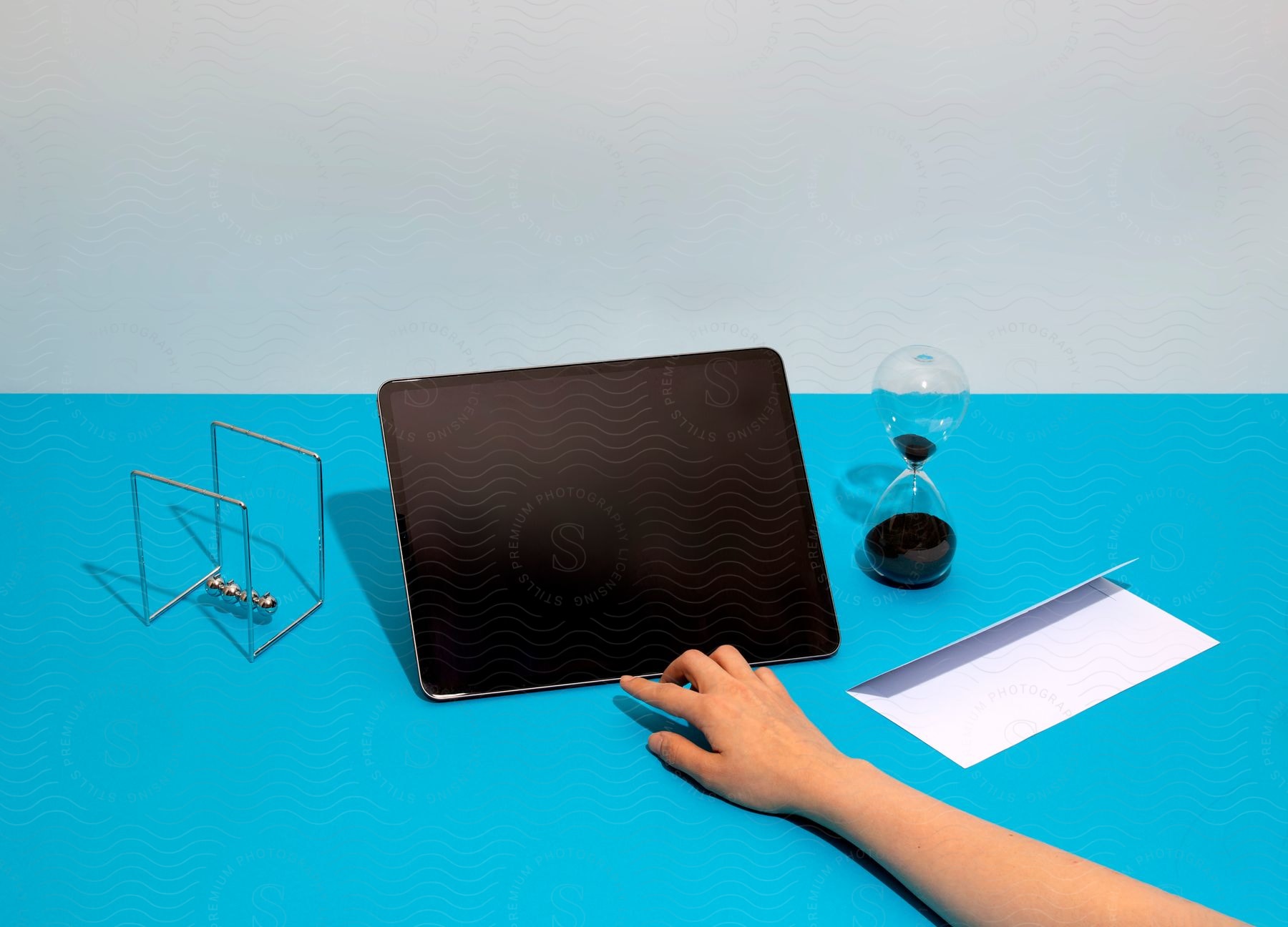 A girl's hand is pointing at a tablet computer on a light blue table between a Newton's cradle and an hourglass